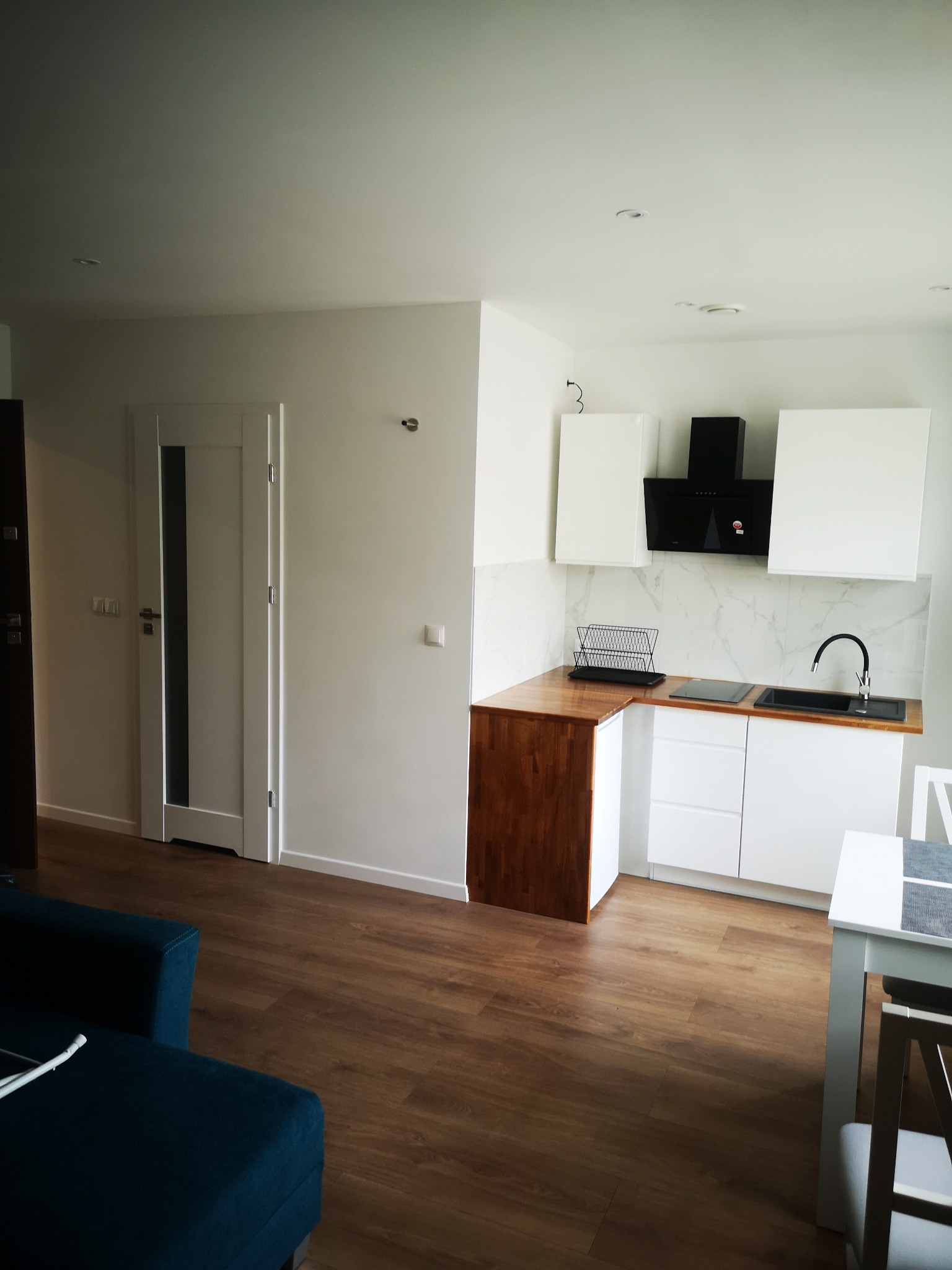 Nowoczesna, jasna kuchnia z białymi szafkami i drewnianymi blatami, czarną armaturą i okapem, widok na salon z ciemnozieloną sofą i drewnianą podłogą.