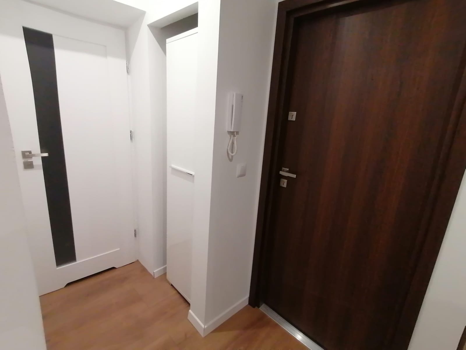 Wnętrze przedpokoju z dwiema parami drzwi: białe drzwi z czarnym paskiem oraz ciemnobrązowe drzwi z metalowymi okuciami, widoczny parkiet i domofon na ścianie.