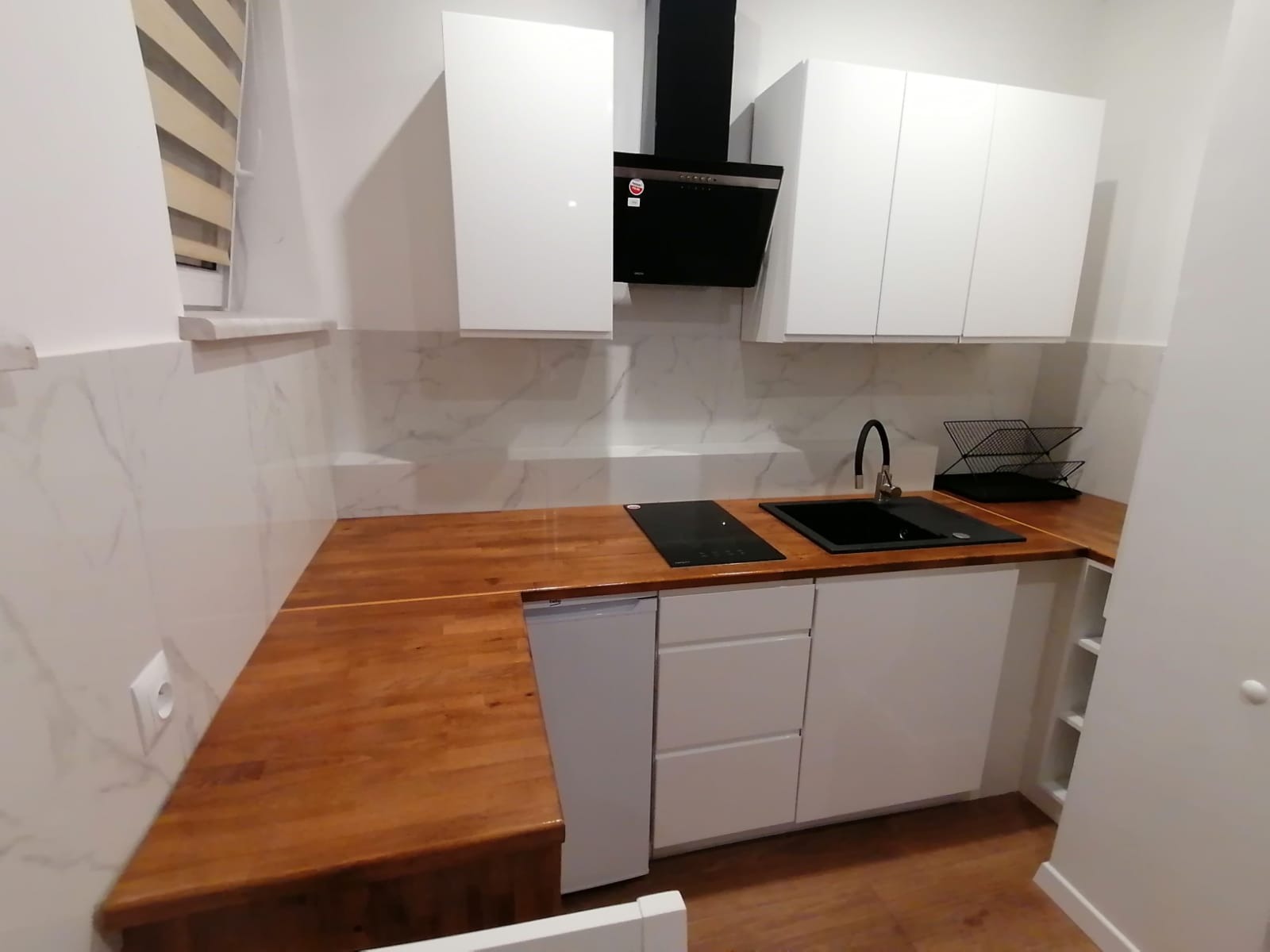 Nowoczesna kuchnia z białymi szafkami, drewnianym blatem i marmurowymi płytkami na ścianie. Widoczny czarny okap, płyta indukcyjna i zlewozmywak. Na blacie suszarka do naczyń.