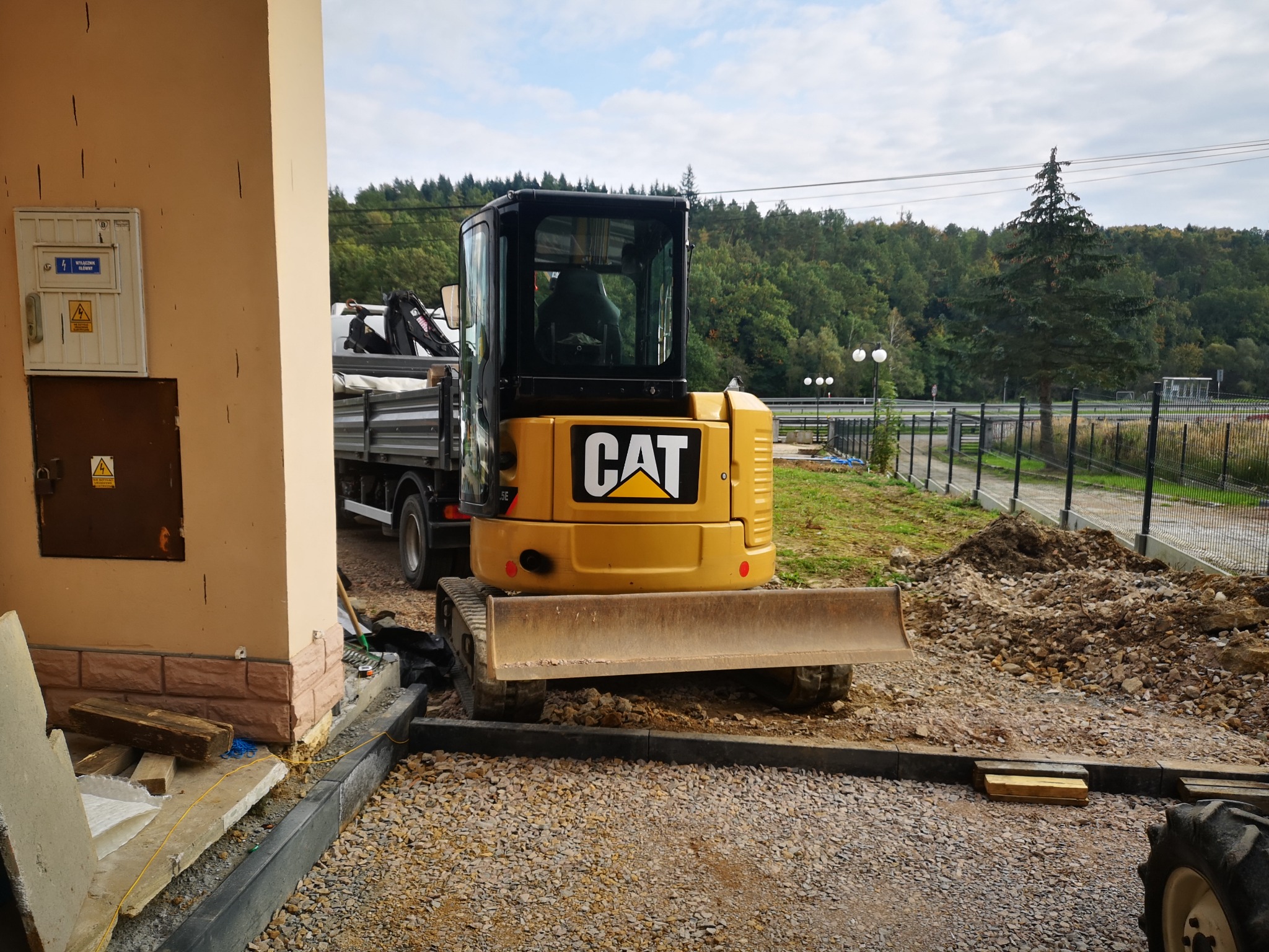 Żółta mini koparka CAT na placu budowy, widok z tyłu, z transportującą ją ciężarówką w tle, obok budynku z ceglaną podmurówką i skrzynką elektryczną.
