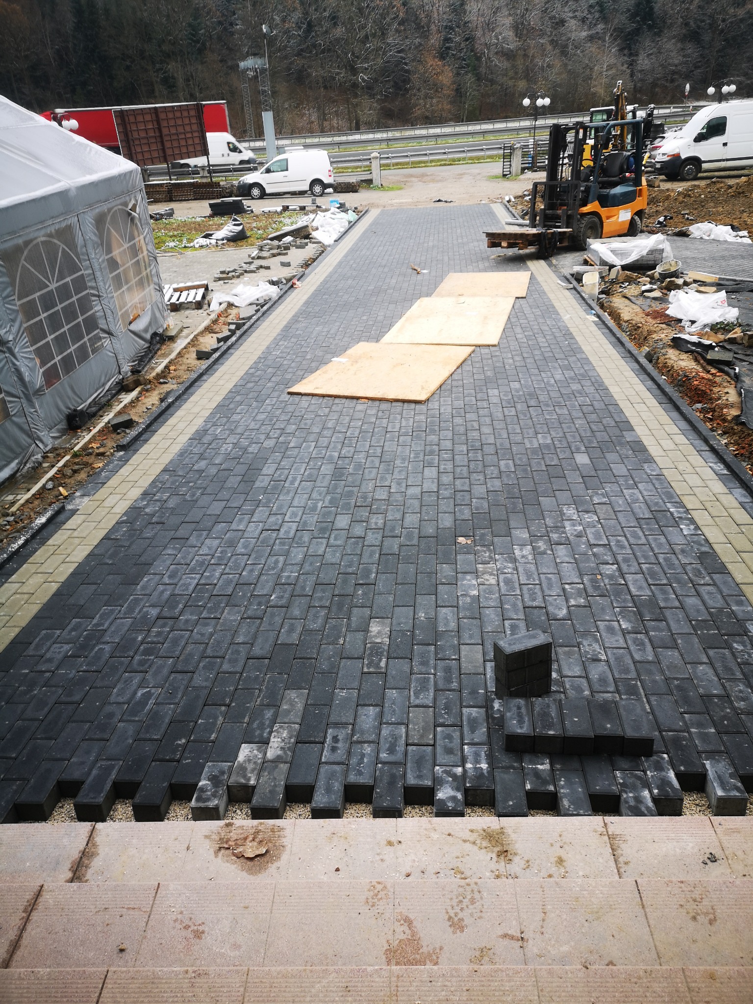 Układanie ciemnoszarej kostki brukowej z jasną obwódką na podjeździe, widoczny wózek widłowy, palety i przygotowane podłoże.