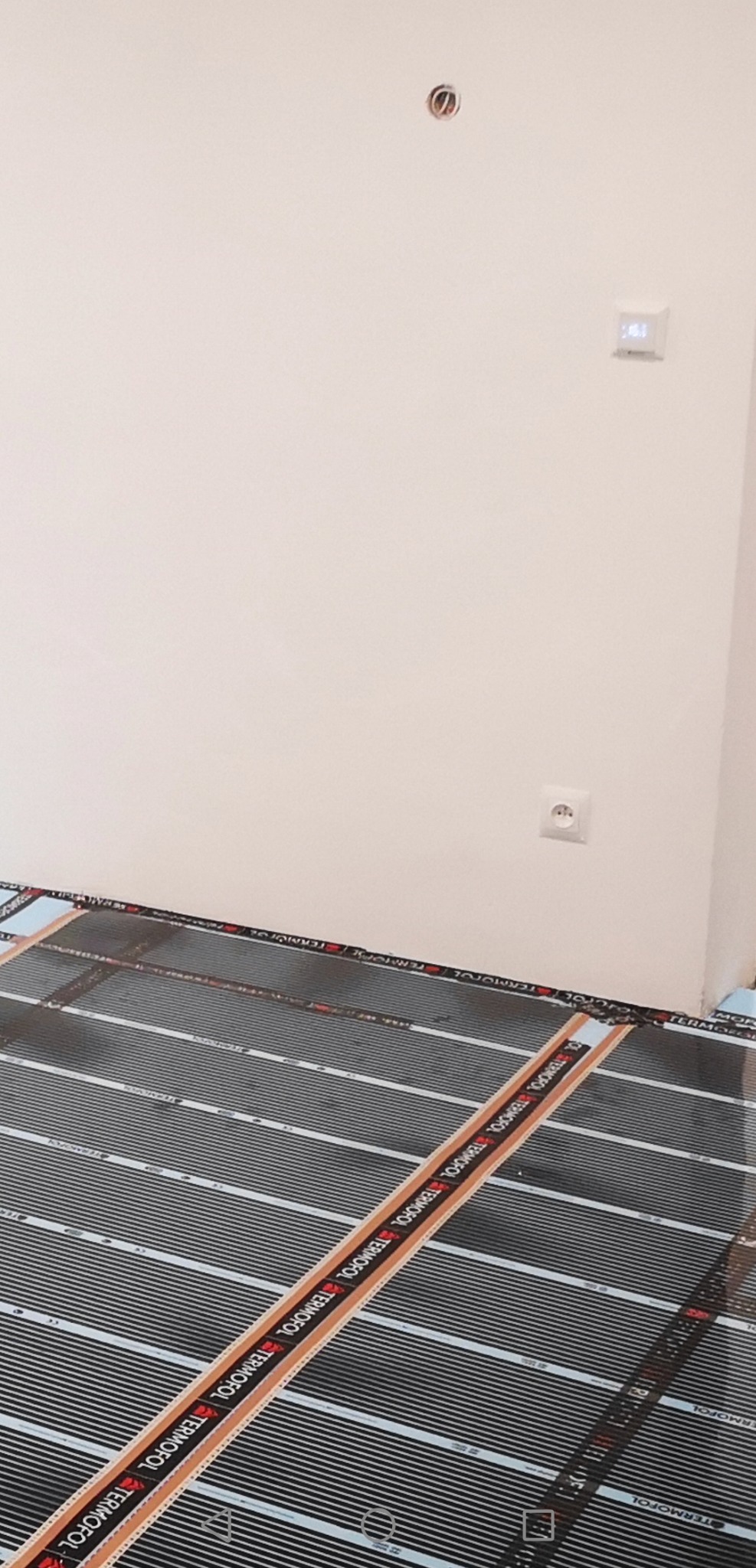 Rozłożona mata grzewcza Termofol na podłodze w pomieszczeniu z gładkimi, białymi ścianami, widoczne gniazdko elektryczne i panel sterowania ogrzewaniem.