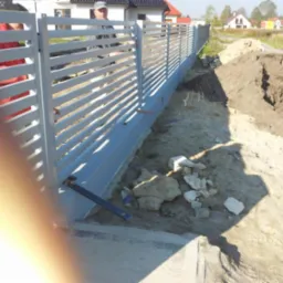 Nowoczesne, poziome ogrodzenie panelowe w kolorze szarym. Widoczny fragment fundamentu i teren wokół. W tle domy jednorodzinne i trawa.