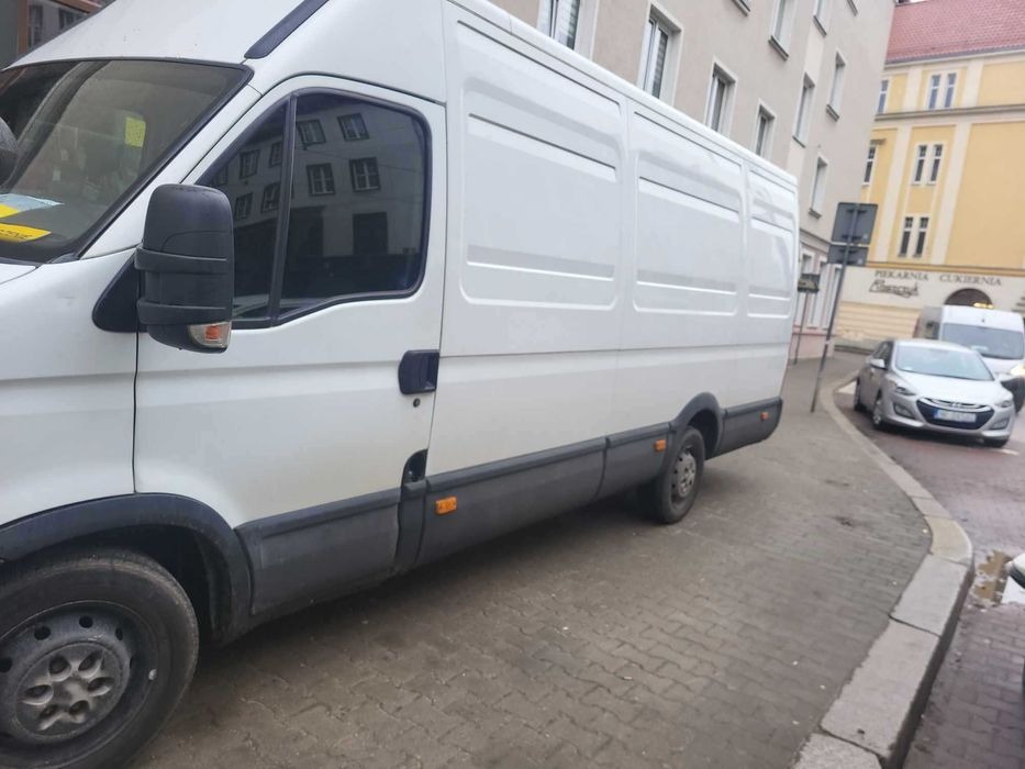 Biały furgon dostawczy zaparkowany na chodniku z kostki brukowej, w tle budynki miejskie i inny pojazd.