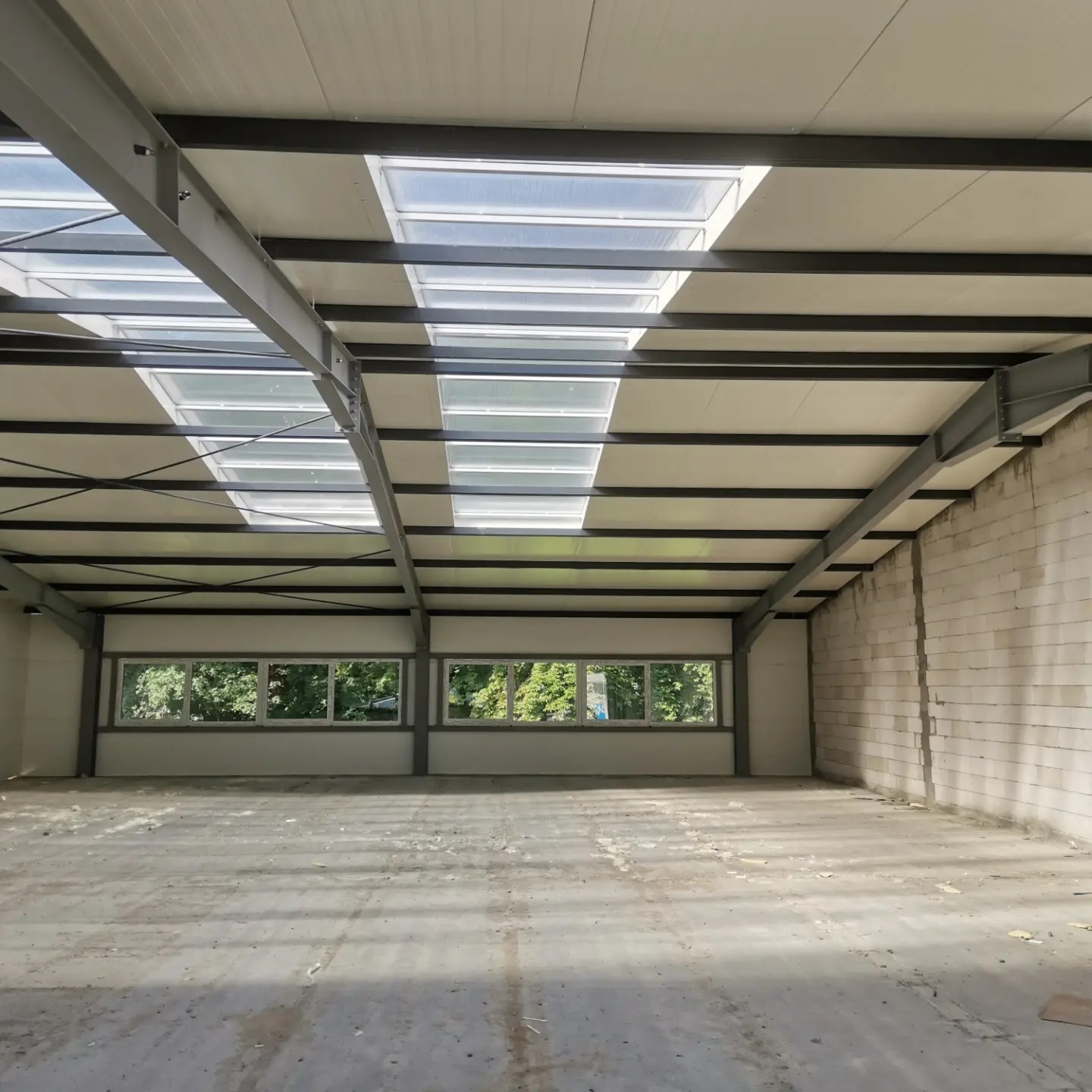 Wnętrze hali w stanie surowym: widoczne stalowe belki konstrukcyjne, okna z widokiem na zieleń, surowy beton na podłodze i ściany z bloczków betonowych.