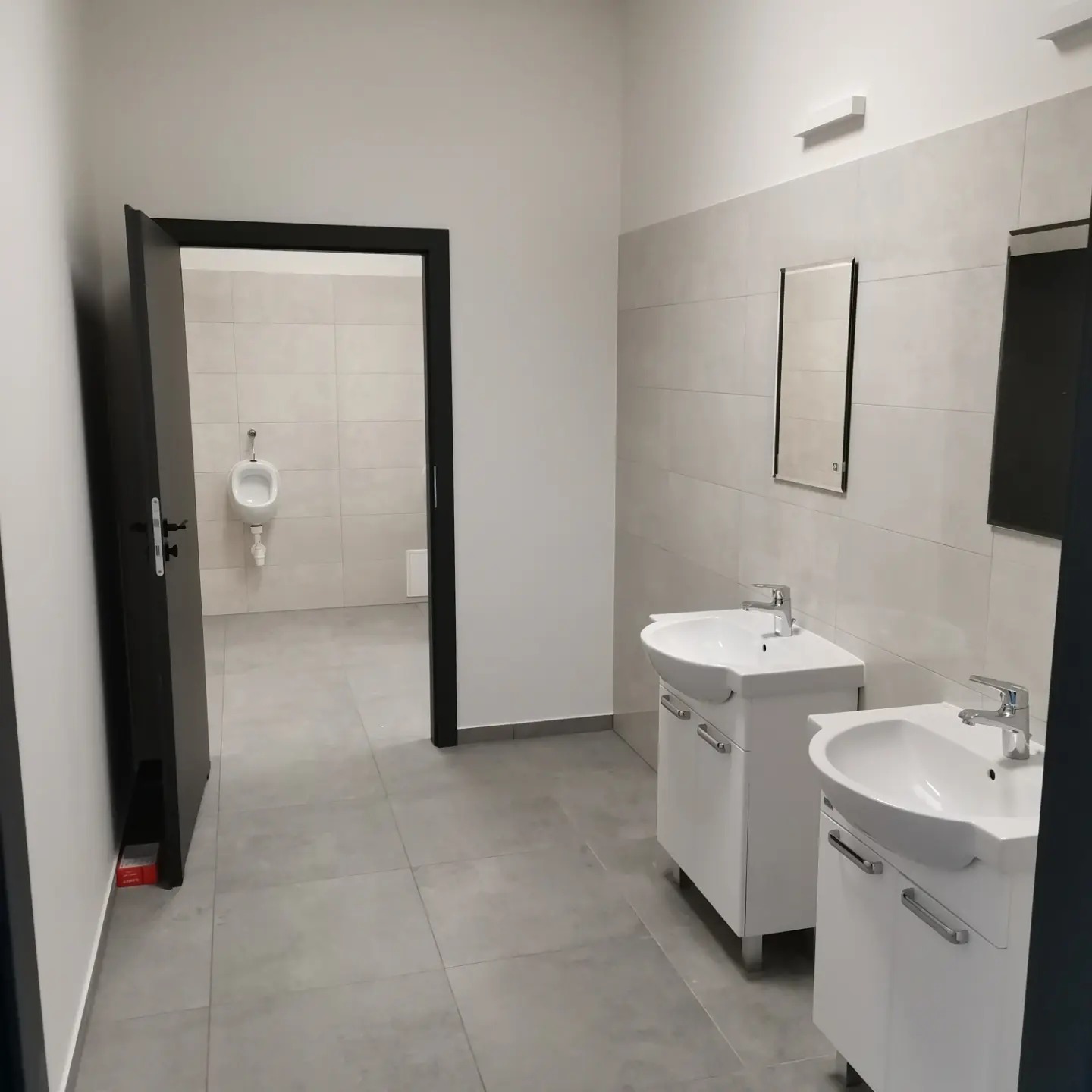 Wnętrze wykończonej łazienki publicznej z dwoma białymi umywalkami na szafkach, lustrem na ścianie z szarych płytek i widocznym pisuar w oddali.