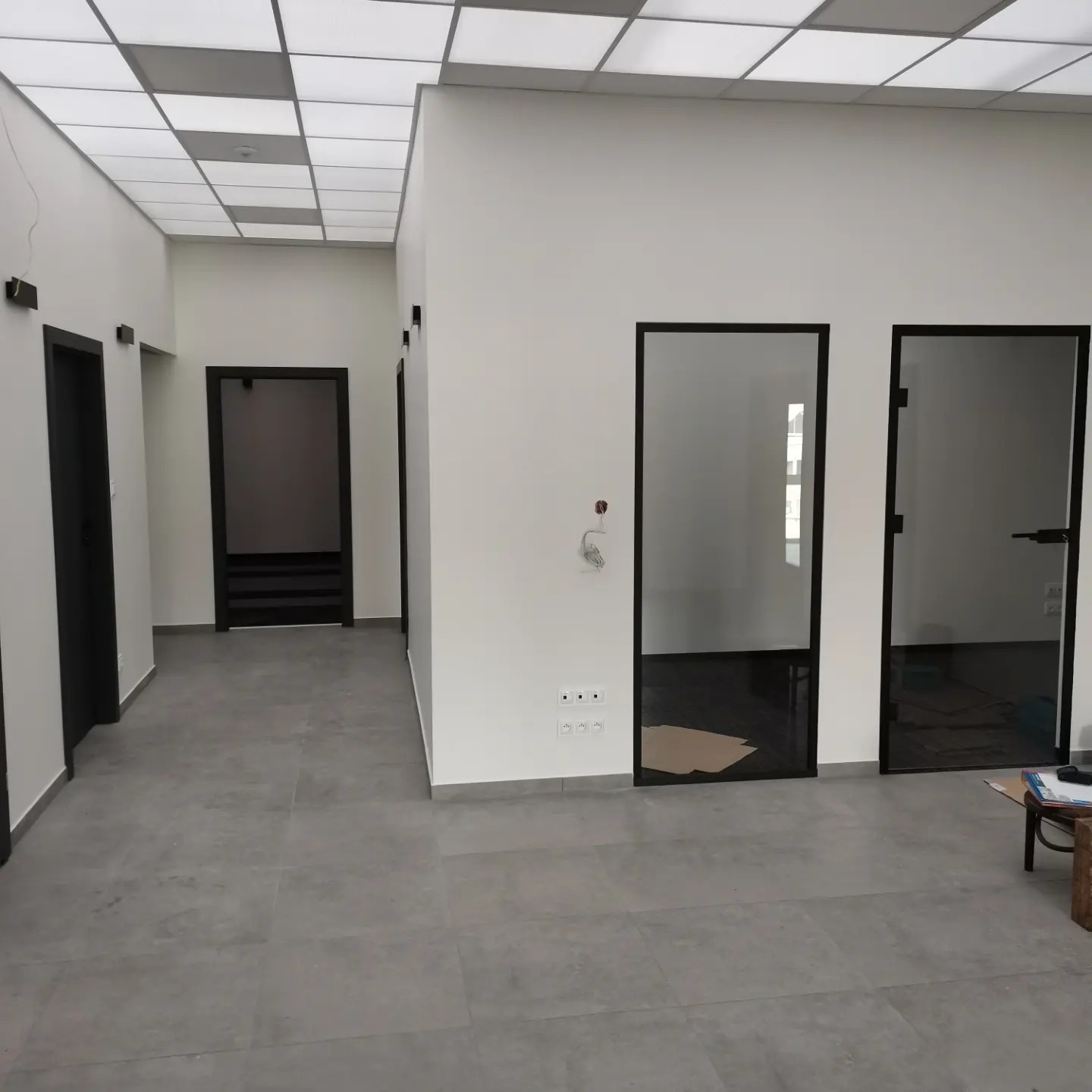 Wnętrze biura z szarymi płytkami podłogowymi, białymi ścianami, czarnymi futrynami drzwi, przeszklonymi drzwiami i kasetonowym oświetleniem sufitowym.