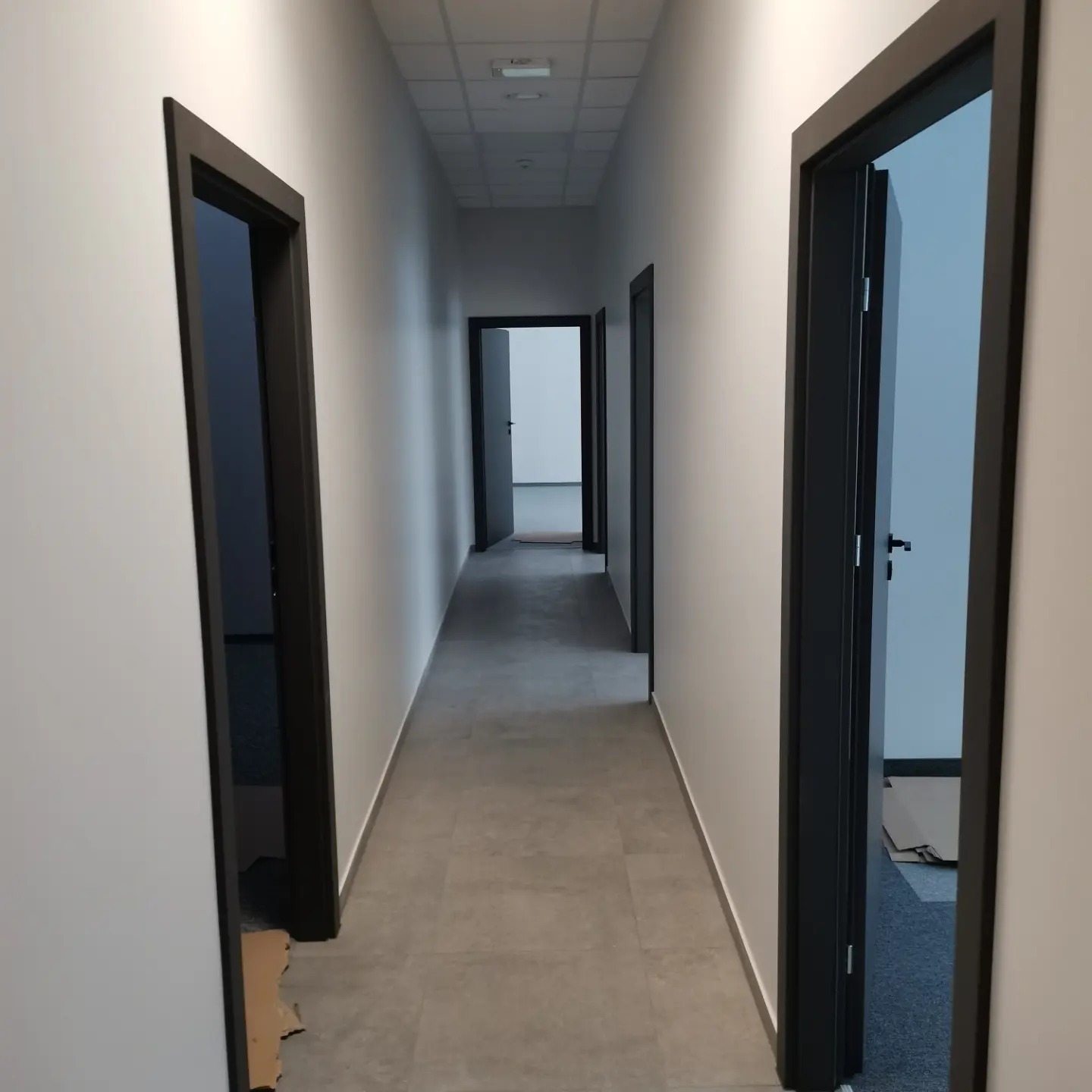 Długi korytarz biurowy z szarymi płytkami podłogowymi i białymi ścianami, widoczne czarne ościeżnice drzwiowe, w tle otwarte drzwi.