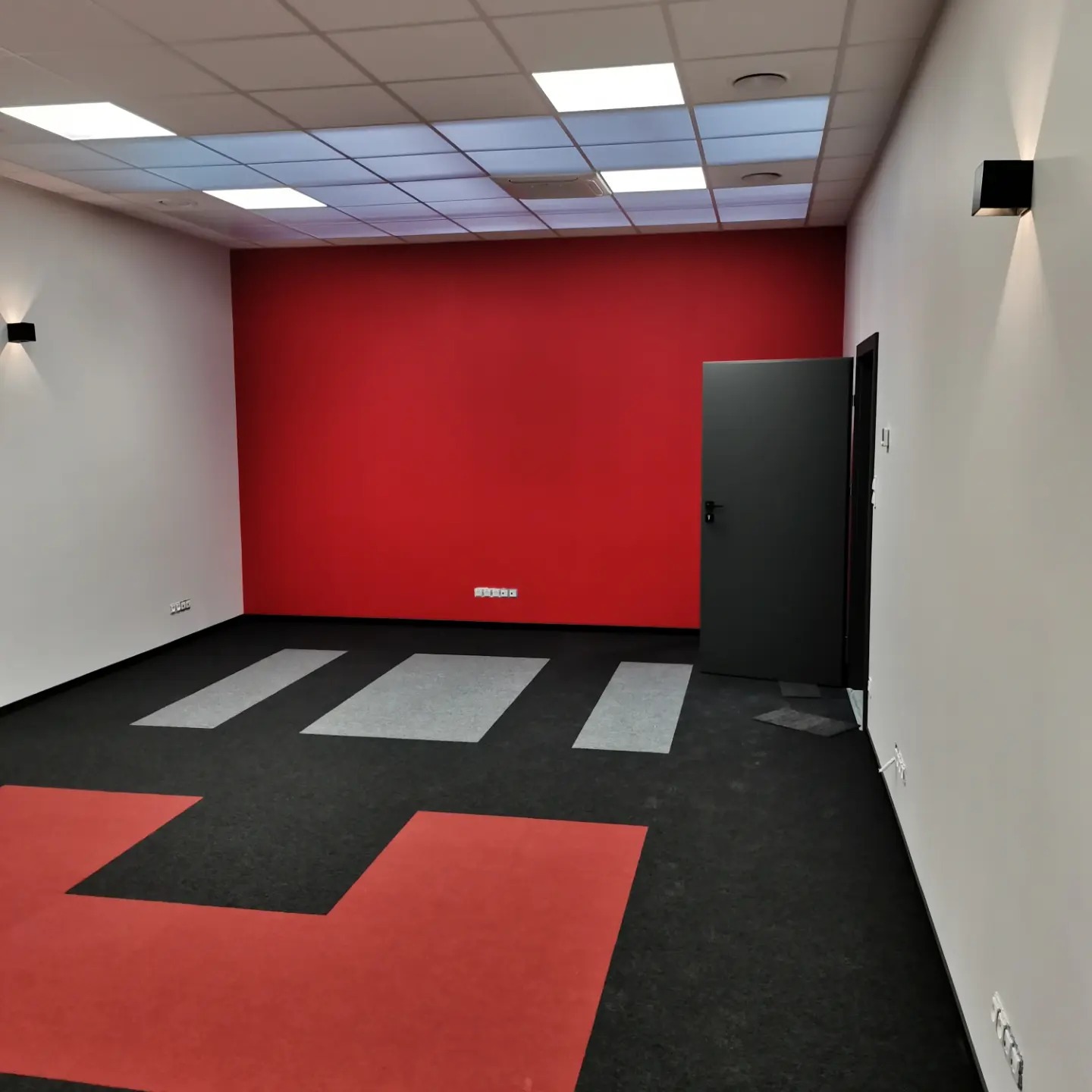 Pomalowane na czerwono ściany w biurze z czarną wykładziną z czerwonymi i szarymi wstawkami, czarne lampy ścienne i drzwi.