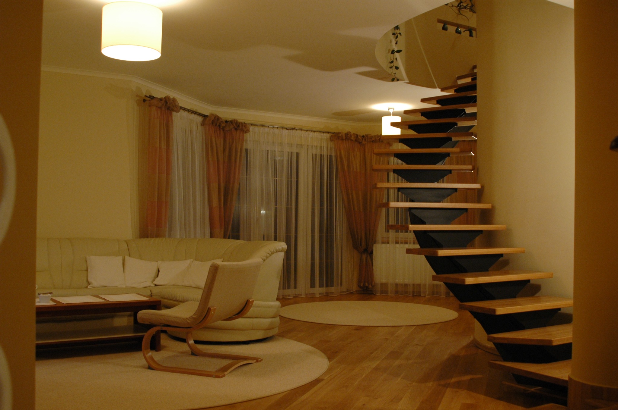 Spiralne schody z drewnianymi stopniami i czarną konstrukcją w przestronnym salonie z parkietem i kremową sofą.