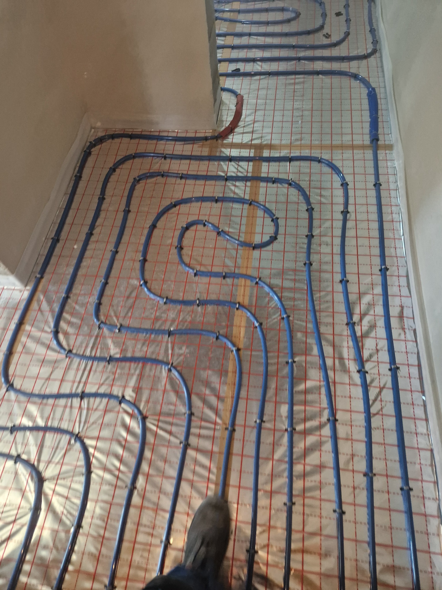 Niebieskie rurki instalacji grzewczej ułożone w pętle na folii z czerwoną siatką, widok z góry, fragment buta w rogu kadru.