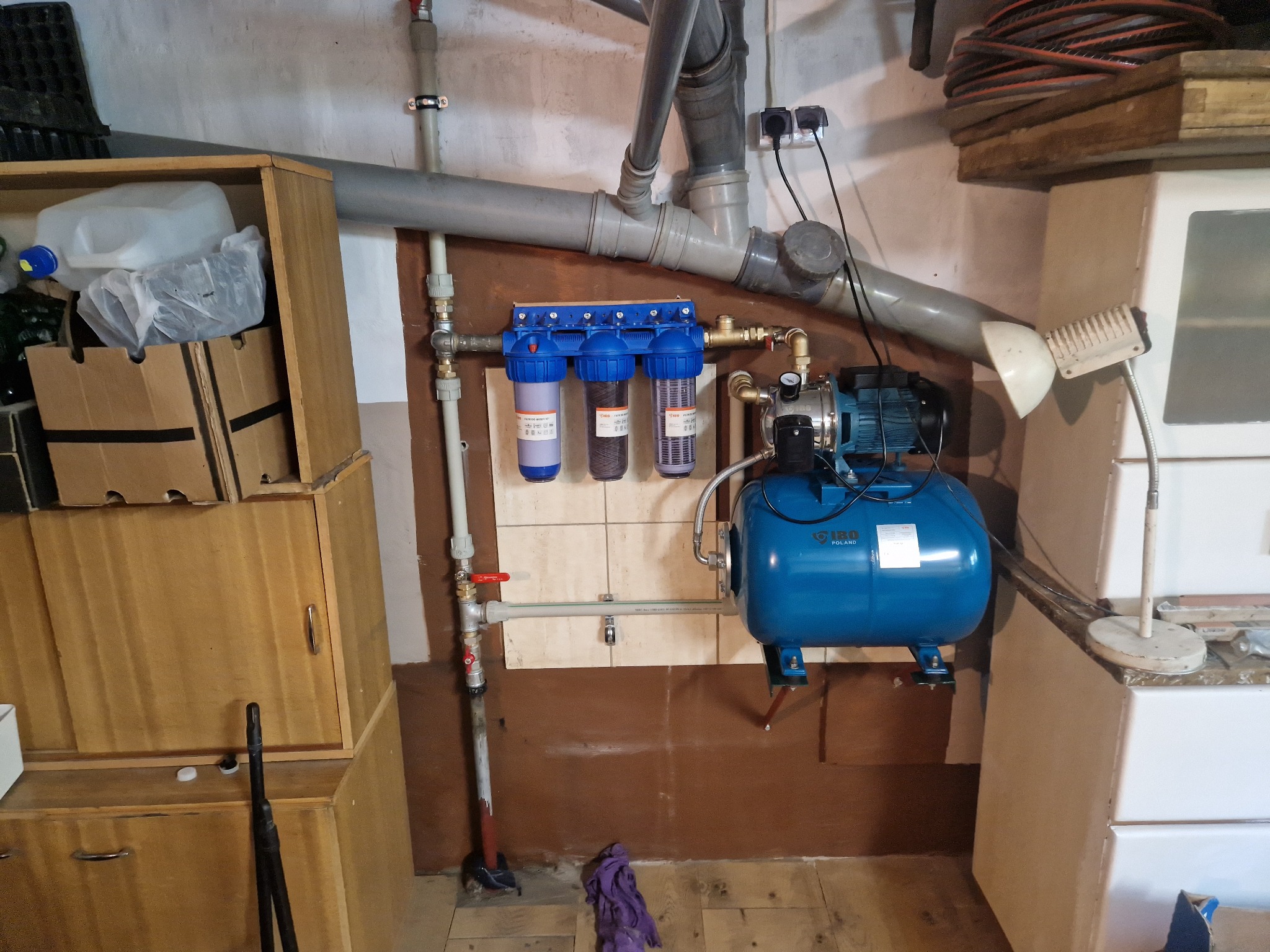 Instalacja hydroforowa z filtrami wody, rurami PCV i armaturą, zamontowana w piwnicy na tle boazerii i szafek.