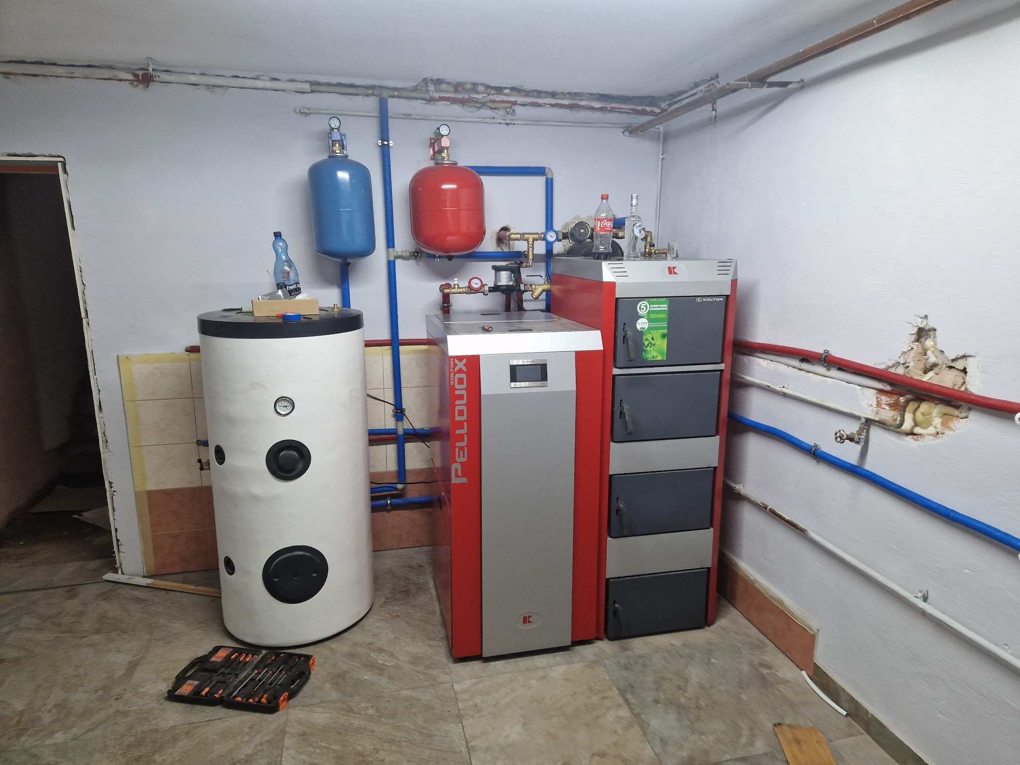 Kotłownia z piecem na pellet, zbiornikiem buforowym i instalacją hydrauliczną, widoczne narzędzia w skrzynce. Nierówności na ścianie i rury instalacji grzewczej.