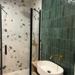 Nowoczesna łazienka z kabiną prysznicową z czarną ramą, ścianą z płytek imitujących lastryko, zielonymi, pionowo ułożonymi płytkami, owalną białą umywalką nablatową na drewnianej półce i złotą...