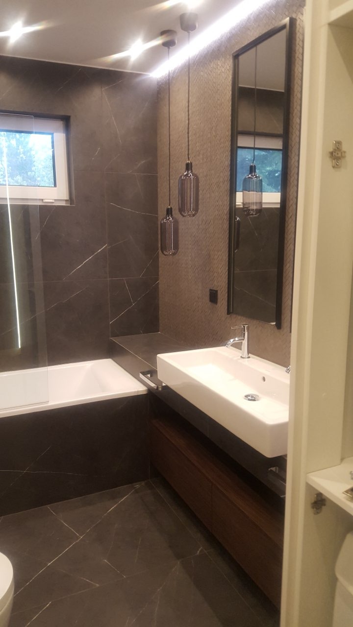 Nowoczesna łazienka z ciemnymi płytkami imitującymi marmur, podwieszana umywalka prostokątna na szafce z ciemnego drewna, wiszące lampy w stylu industrialnym i podświetlane lustro.