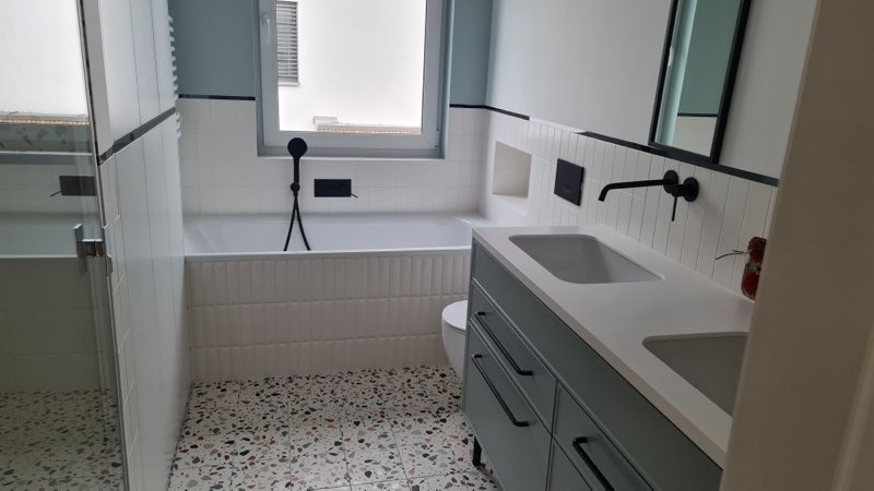 Nowoczesna łazienka z biało-szarą kolorystyką, mozaikową podłogą, wanną obudowaną pionowymi płytkami, czarną armaturą i szafką pod umywalką w odcieniu szaro-zielonym.