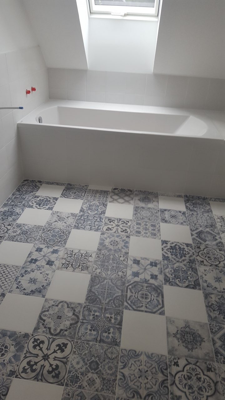 Biała wanna w łazience na poddaszu z oknem dachowym, wykończona białą glazurą i podłogą z patchworkowych płytek w odcieniach bieli i niebieskiego.