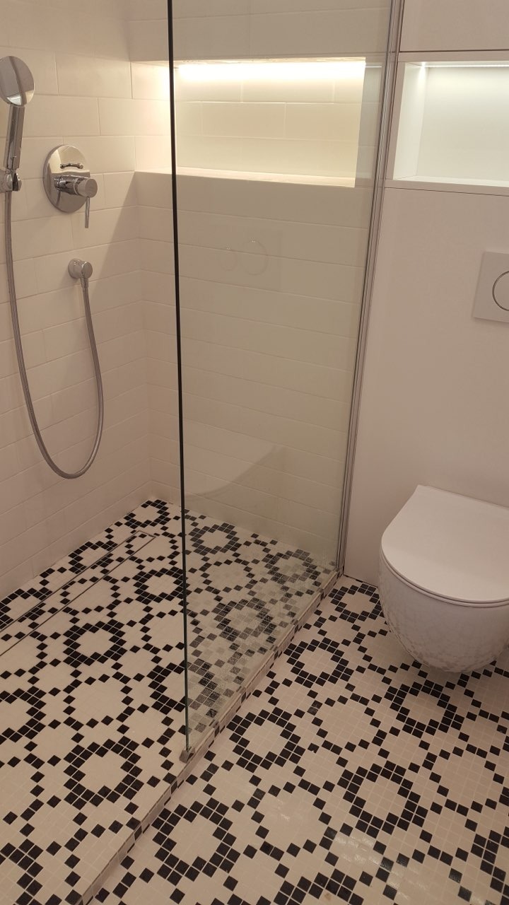 Nowoczesna łazienka z kabiną prysznicową typu walk-in, wykończona białą glazurą i czarno-białą mozaiką podłogową o geometrycznym wzorze, z widocznym zestawem prysznicowym i podświetlaną wnęką ścienną.