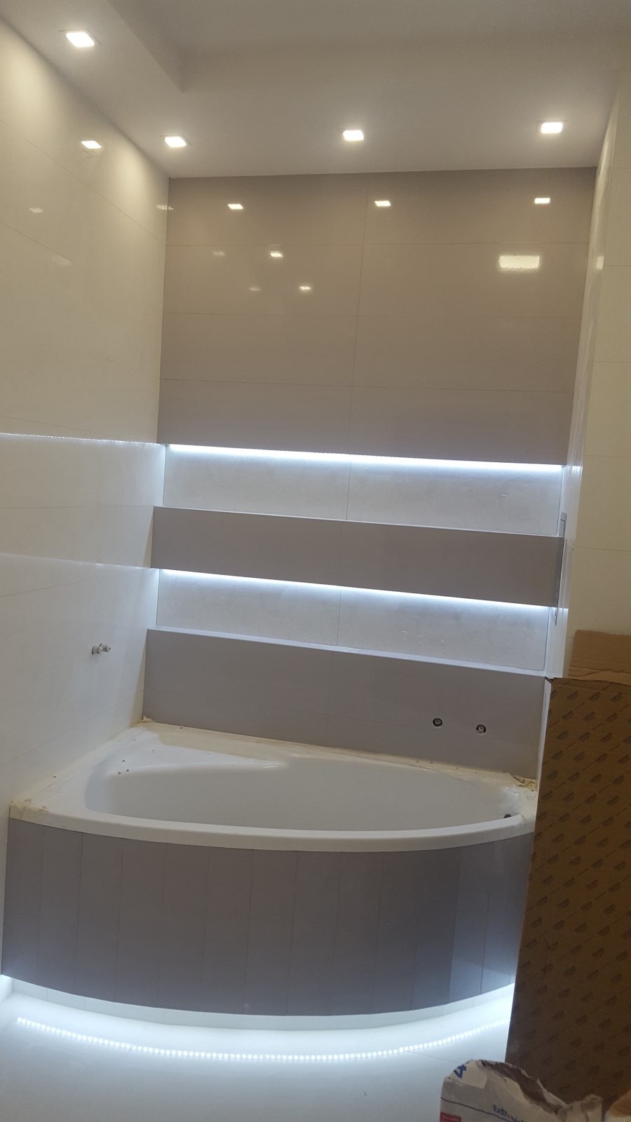 Nowoczesna łazienka z narożną wanną obudowaną szarymi panelami, podświetlona listwami LED umieszczonymi pod wanną i na półkach ściennych, białe płytki na ścianach i punktowe oświetlenie sufitowe.