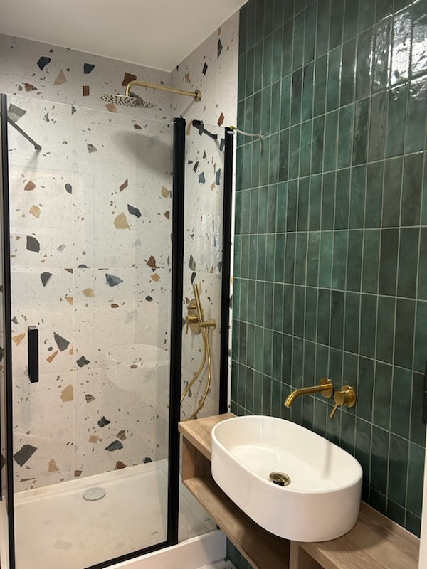 Nowoczesna łazienka z kabiną prysznicową z czarną ramą, ścianą z płytek imitujących lastryko, zielonymi, pionowo ułożonymi płytkami, owalną białą umywalką nablatową na drewnianej półce i złotą...