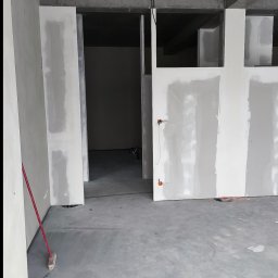 „DARO” Firma Remontowo - Wykończeniowa Dariusz Bąk - Surowy stan wnętrza z płytami gipsowo-kartonowymi, widoczne kable i betonowy sufit. Czerwona miotła oparta o ścianę. Wnętrze w trakcie wykańczania.