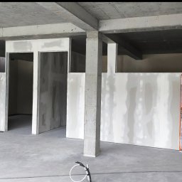 „DARO” Firma Remontowo - Wykończeniowa Dariusz Bąk - Surowy stan wnętrza z płytami gipsowo-kartonowymi, widoczne kable na podłodze, betonowy strop i kolumny. Drzwi zabezpieczone folią. Wnętrze w trakcie prac wykończeniowych.