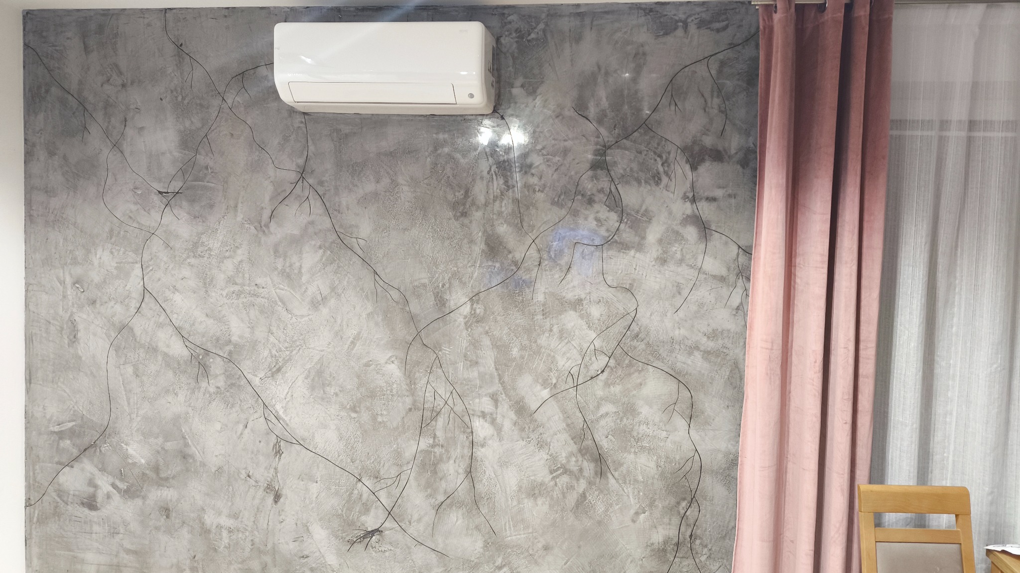Szara ściana z efektem betonu, dekorowana czarnymi, cienkimi liniami przypominającymi gałązki. Na ścianie zamontowany biały klimatyzator. Obok różowe zasłony i fragment krzesła.