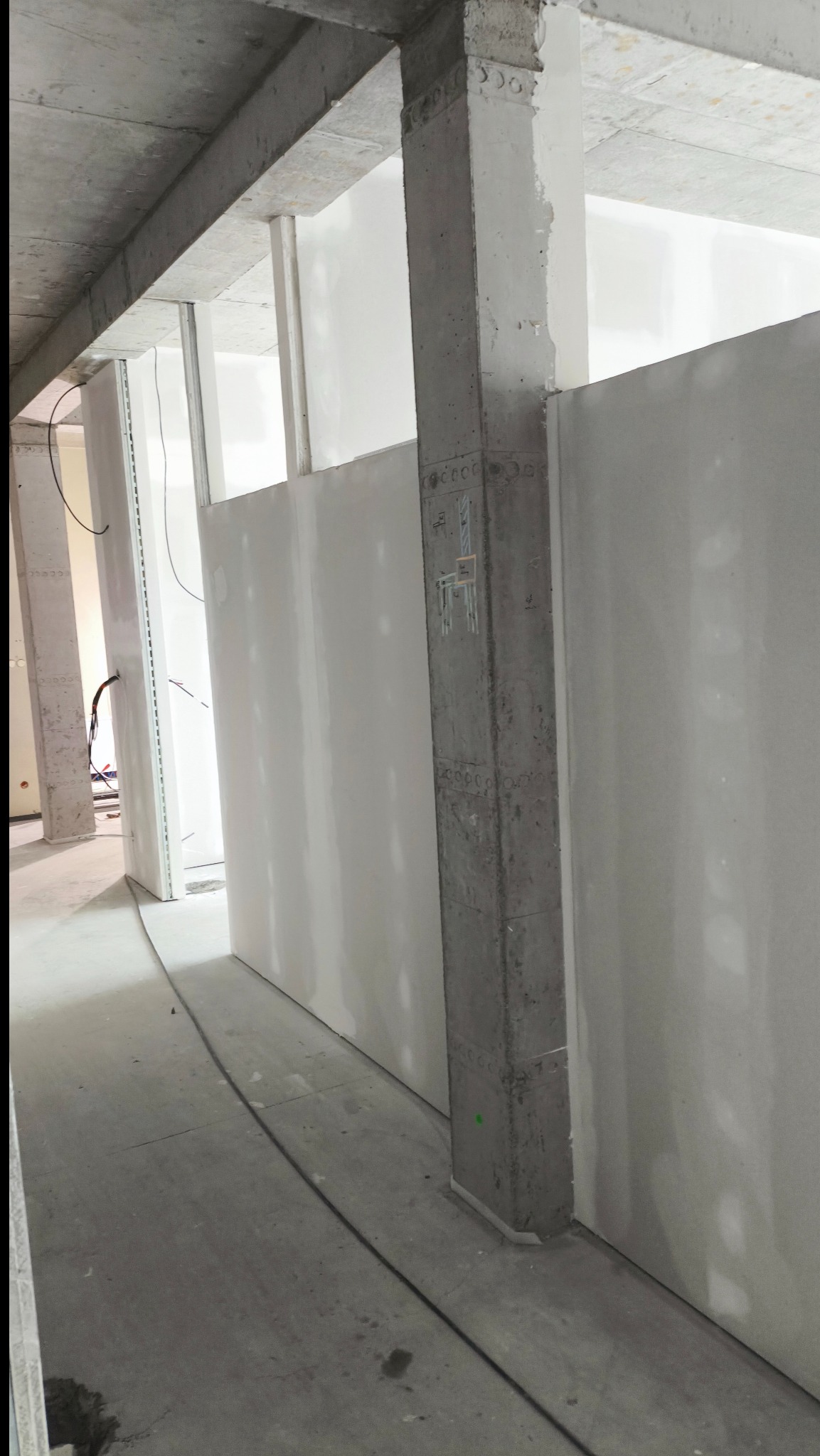Wnętrze w trakcie prac wykończeniowych: surowy betonowy filar na pierwszym planie, ściany z płyt karton-gips, widoczne kable i elementy konstrukcyjne.