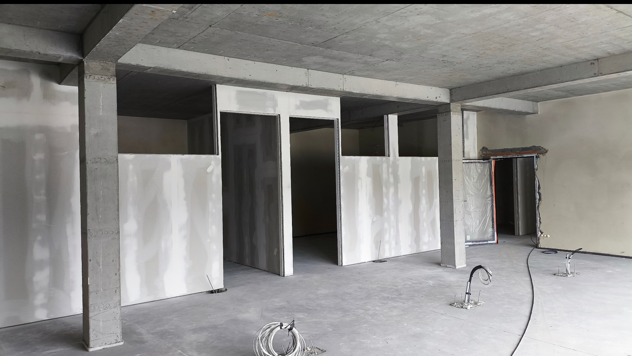 Surowy stan wnętrza z betonowymi kolumnami i ścianami z płyt gipsowo-kartonowych, przygotowany do dalszych prac wykończeniowych. Widoczne instalacje elektryczne.
