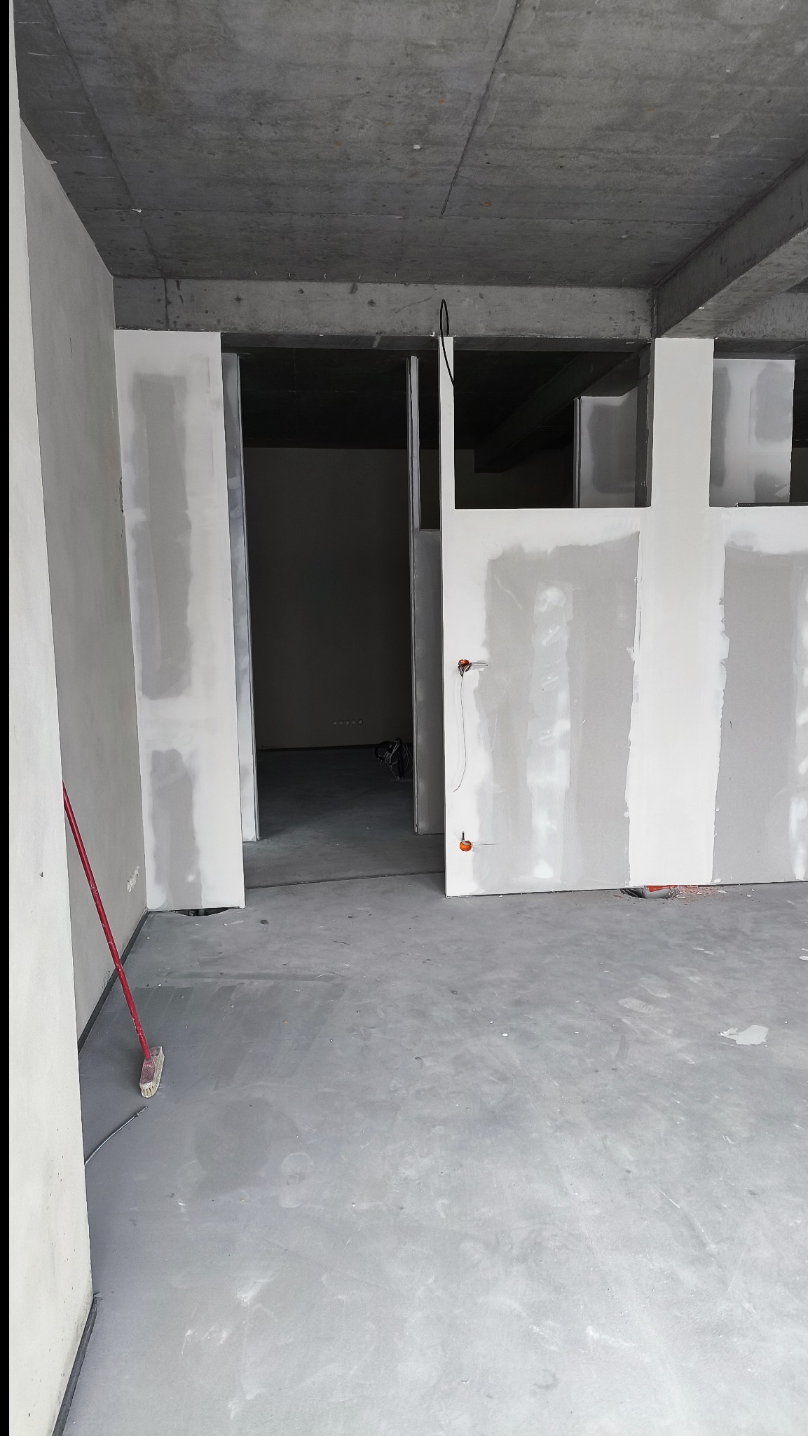 Surowy stan wnętrza z płytami gipsowo-kartonowymi, widoczne kable i betonowy sufit. Czerwona miotła oparta o ścianę. Wnętrze w trakcie wykańczania.