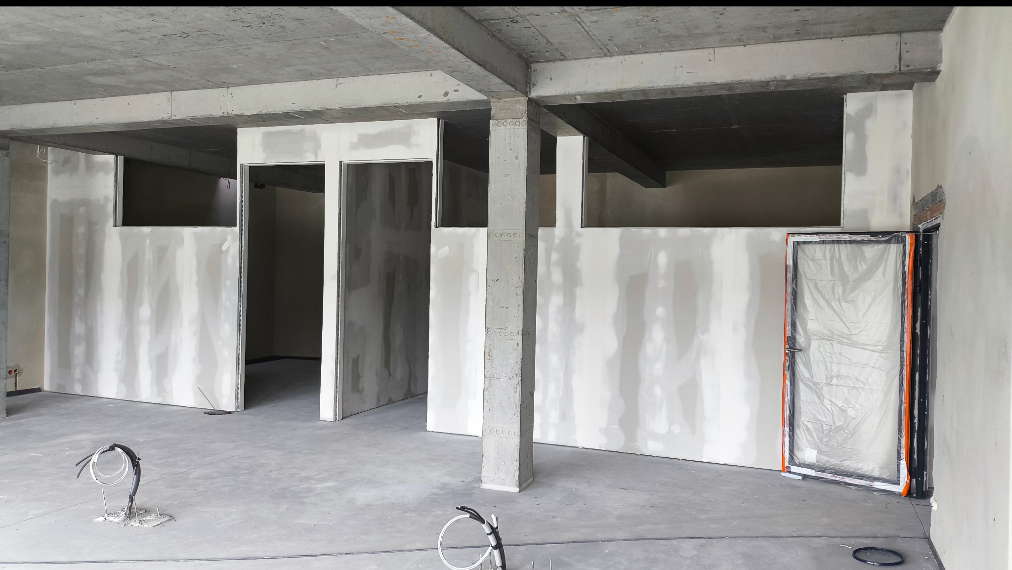 Surowy stan wnętrza z płytami gipsowo-kartonowymi, widoczne kable na podłodze, betonowy strop i kolumny. Drzwi zabezpieczone folią. Wnętrze w trakcie prac wykończeniowych.