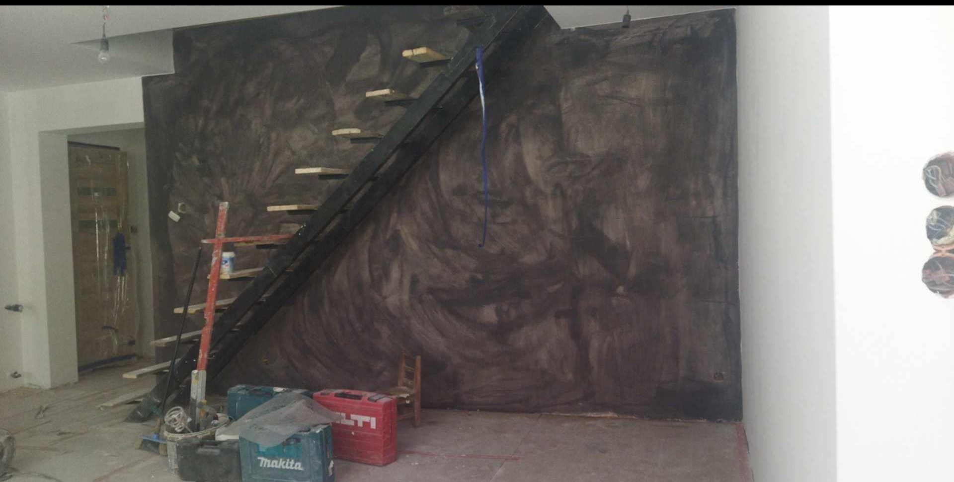 Ściana z efektem dekoracyjnym w odcieniach szarości, z drewnianymi schodami i narzędziami budowlanymi w pomieszczeniu w trakcie wykańczania. Widoczne pędzle i drabina.