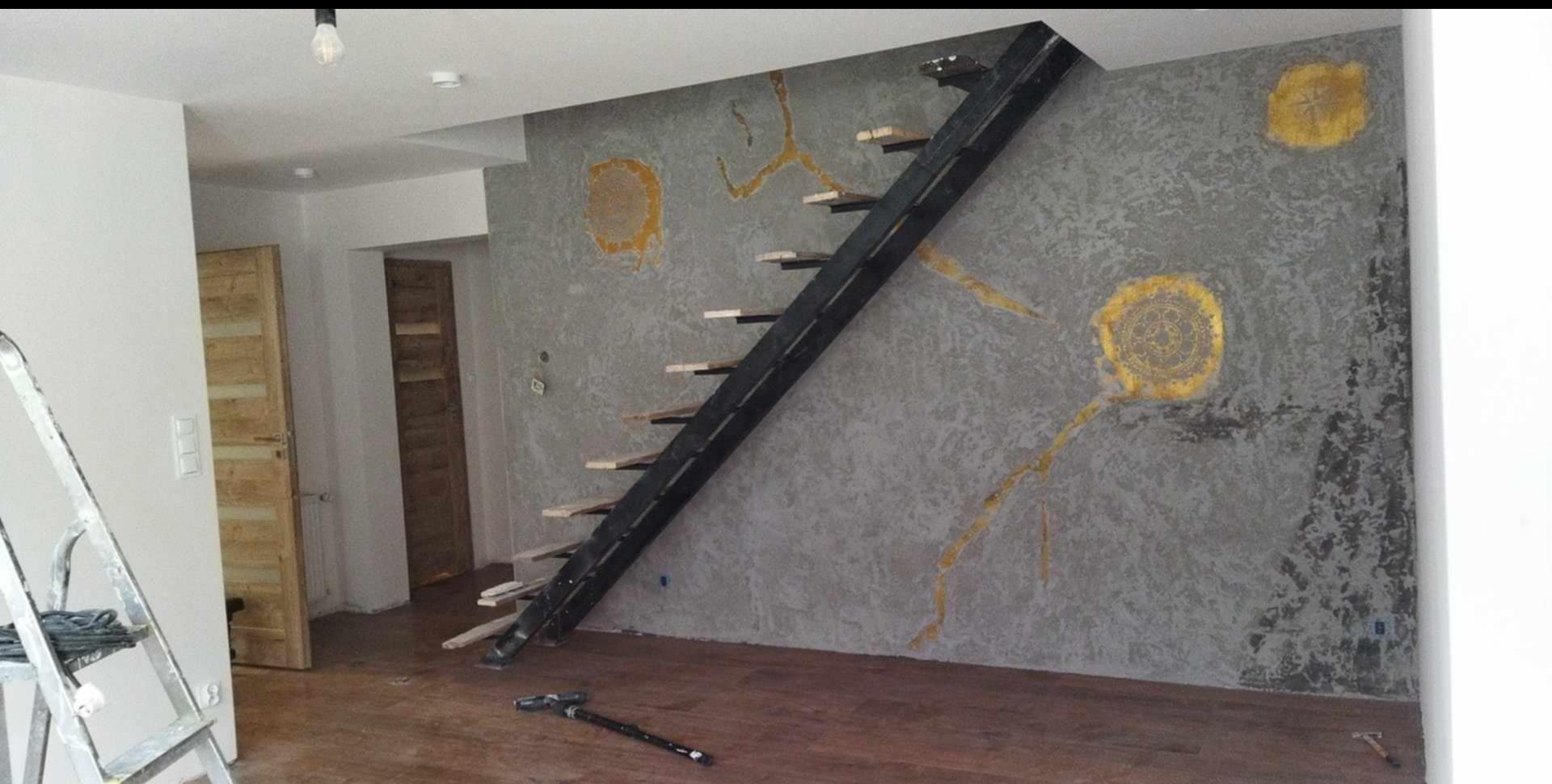 Szara ściana z dekoracyjnymi, złotymi wzorami i czarne schody z drewnianymi stopniami. Widoczne drewniane drzwi i drabina malarska. Podłoga z paneli.
