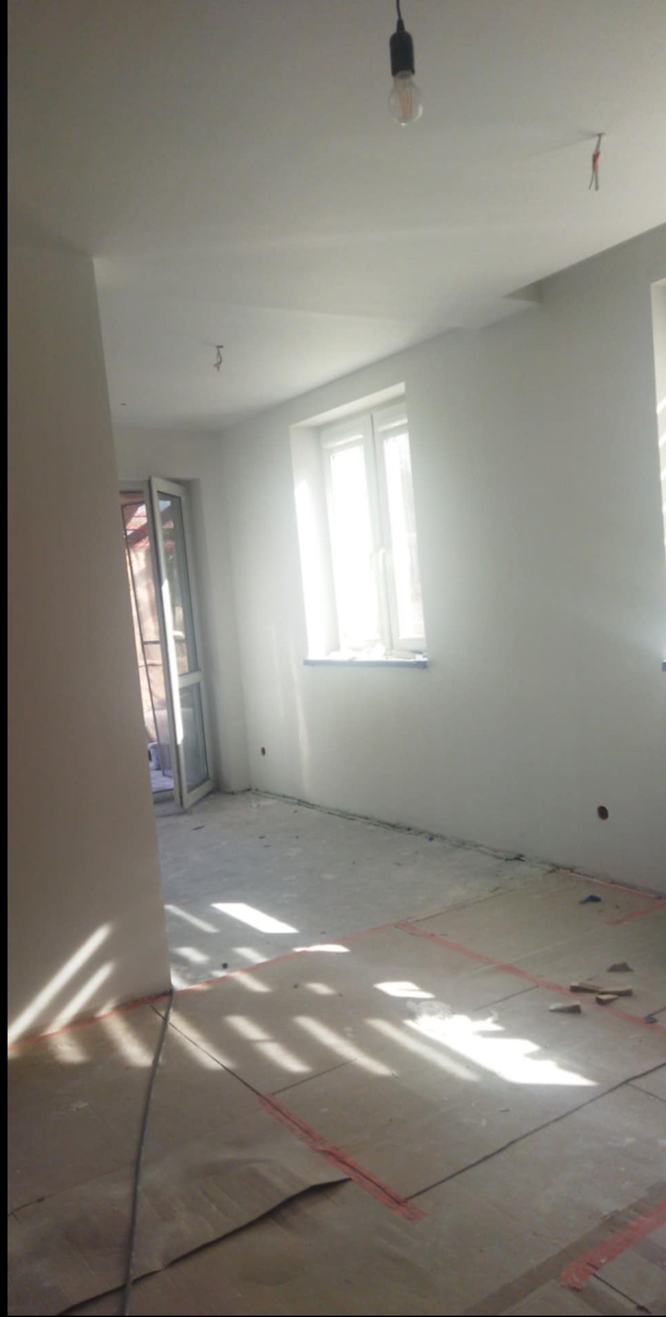Jasne wnętrze w trakcie wykańczania: białe ściany, okno z widokiem na zewnątrz, podłoga zabezpieczona kartonem. Widoczne kable i żarówka na suficie.