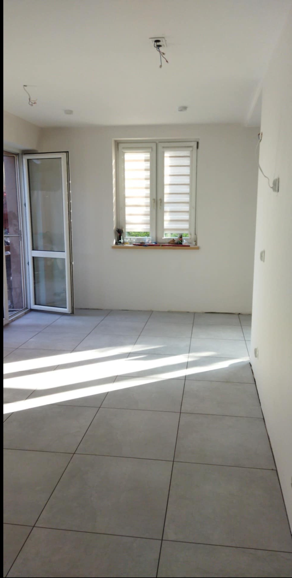 Jasne, kwadratowe płytki podłogowe w nowym pomieszczeniu z oknem i otwartym balkonem. Widoczne niedokończone instalacje elektryczne na suficie. Minimalistyczny styl.