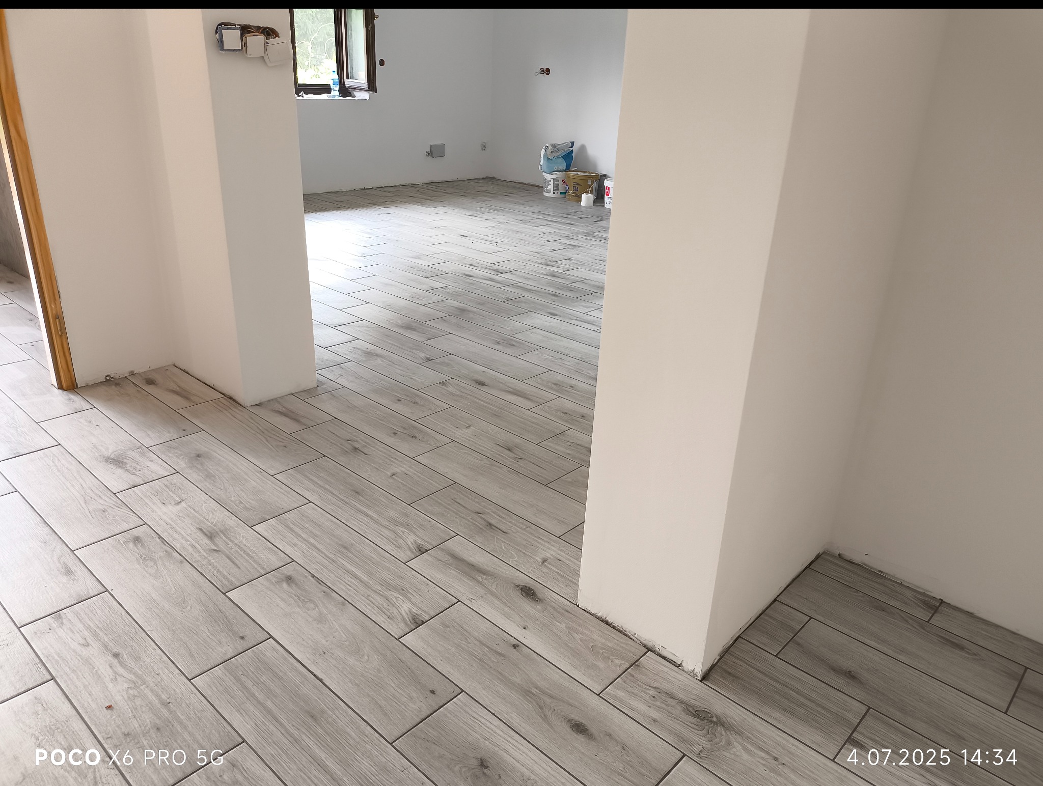 Ułożona podłoga z płytek imitujących drewno w jasnym odcieniu szarości, widok z perspektywy podłogi, z fragmentem białej ściany i otwartym przejściem do innego pomieszczenia.