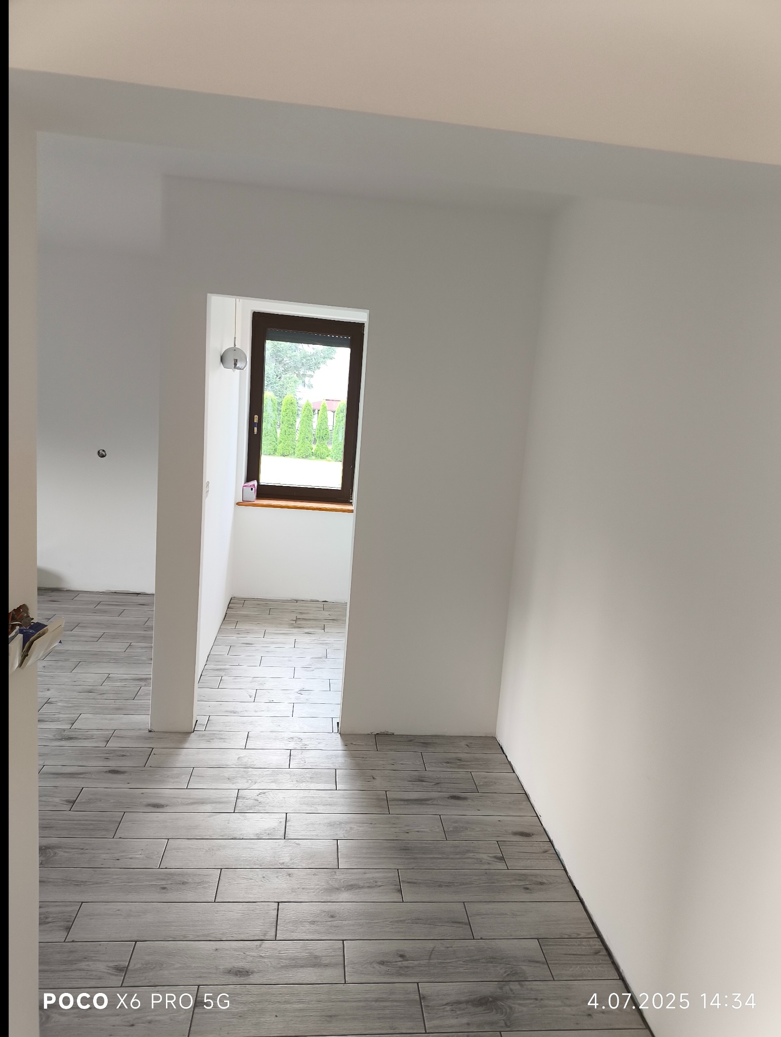 Jasne, drewnopodobne płytki ułożone w korytarzu z widokiem na okno. Świeżo wykończone wnętrze z białymi ścianami i naturalnym światłem.