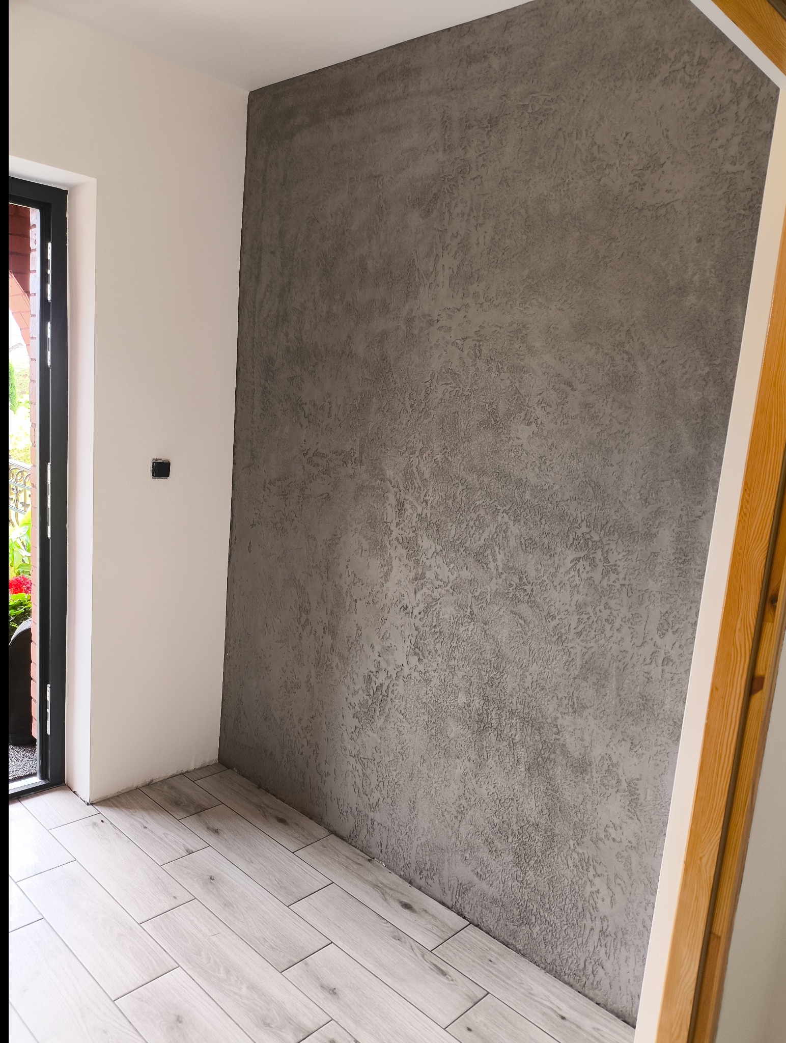 Szara ściana z efektem dekoracyjnym w nowym wnętrzu. Jasne panele podłogowe imitujące drewno. Fragment ramy drzwiowej z naturalnego drewna.