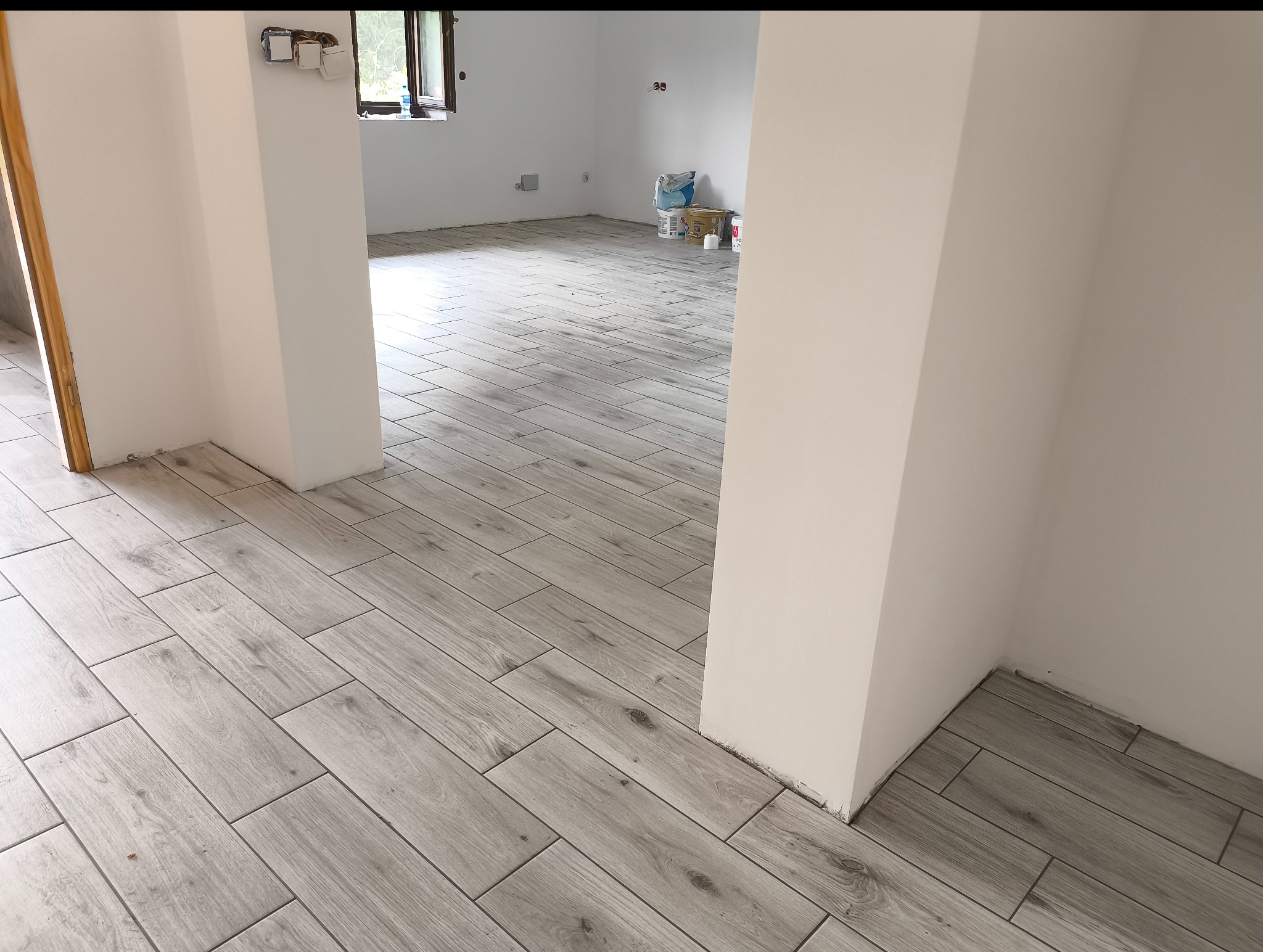 Ułożona podłoga z płytek imitujących drewno w jasnym odcieniu szarości, widok z perspektywy wejścia do pomieszczenia, ściany w kolorze złamanej bieli.