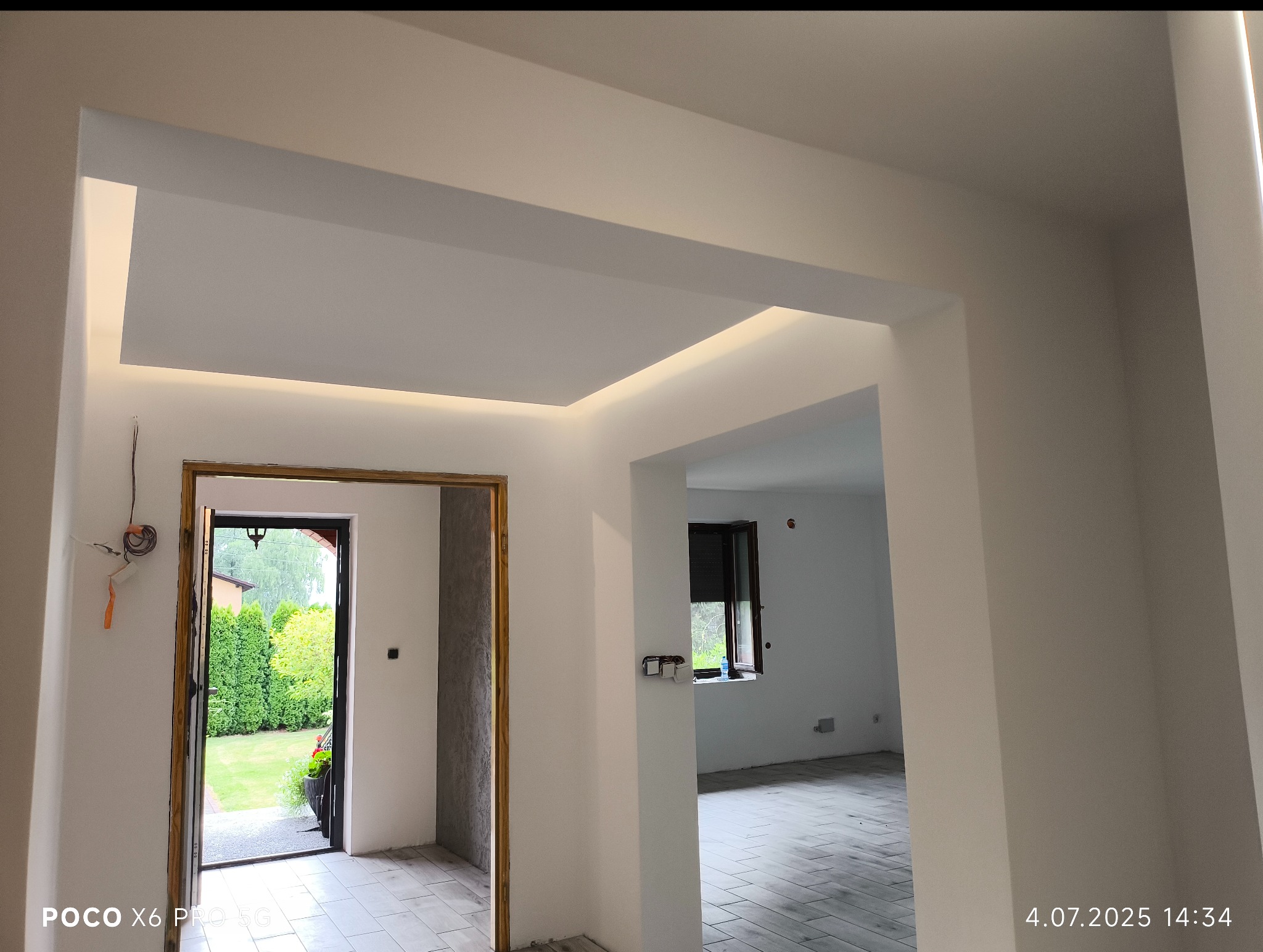 Nowoczesne wykończenie wnętrza: białe ściany, podwieszany sufit z oświetleniem LED, widok na ogród przez otwarte drzwi. Minimalistyczny design, świeżo po remoncie.