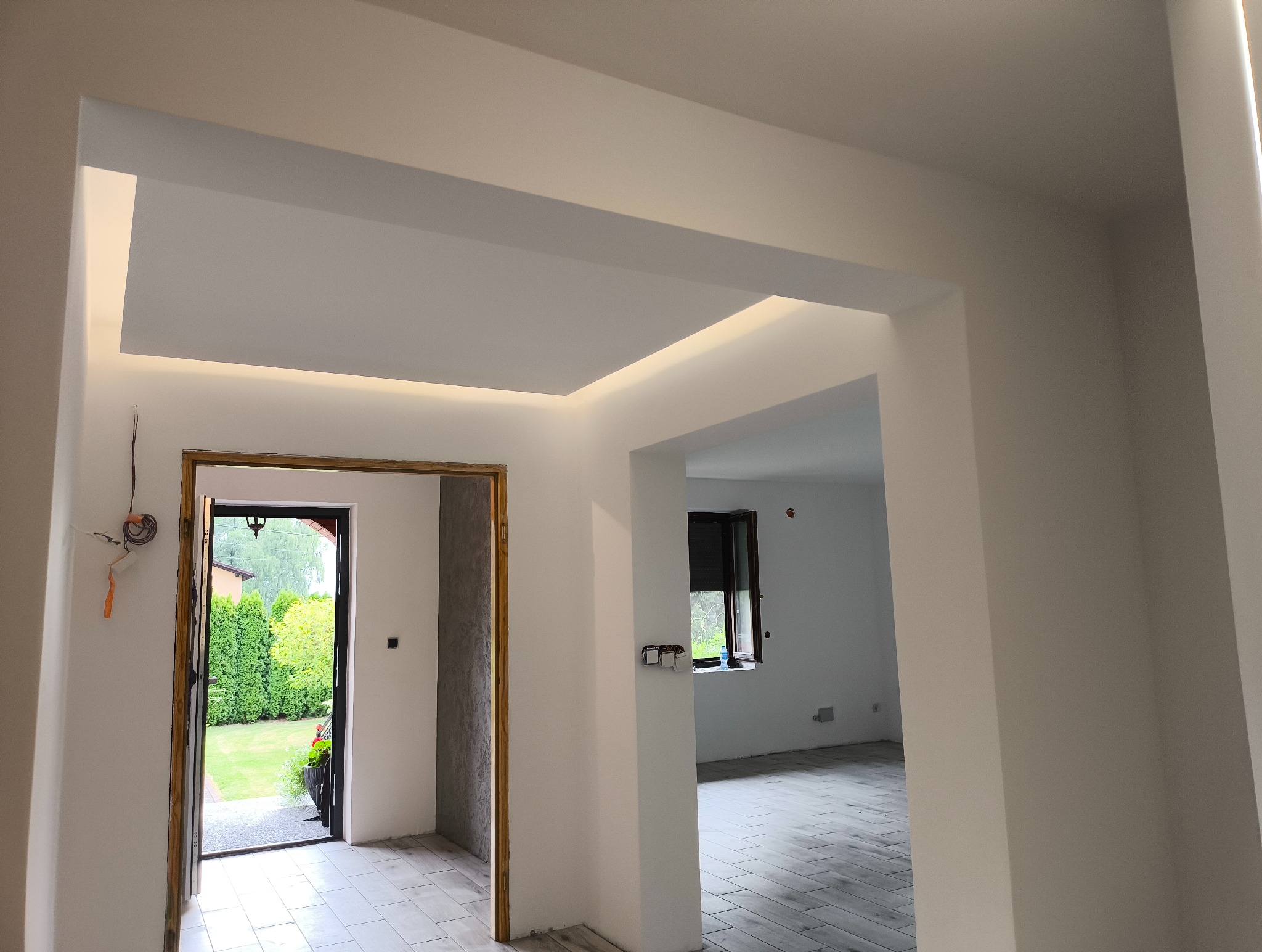 Nowoczesne wykończenie wnętrza: białe ściany, sufit podwieszany z oświetleniem LED, widok na ogród przez otwarte drzwi. Minimalistyczny design, jasne kolory, świeżo po remoncie.