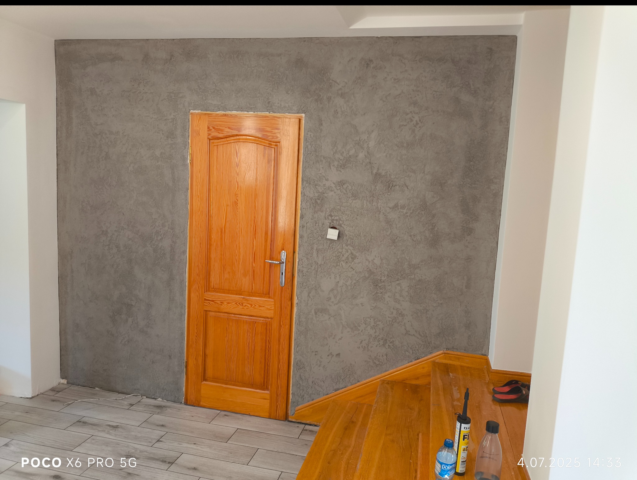 Ściana z efektem dekoracyjnym w odcieniach szarości, drewniane drzwi z ościeżnicą i fragment schodów. Widoczne materiały budowlane na schodach.