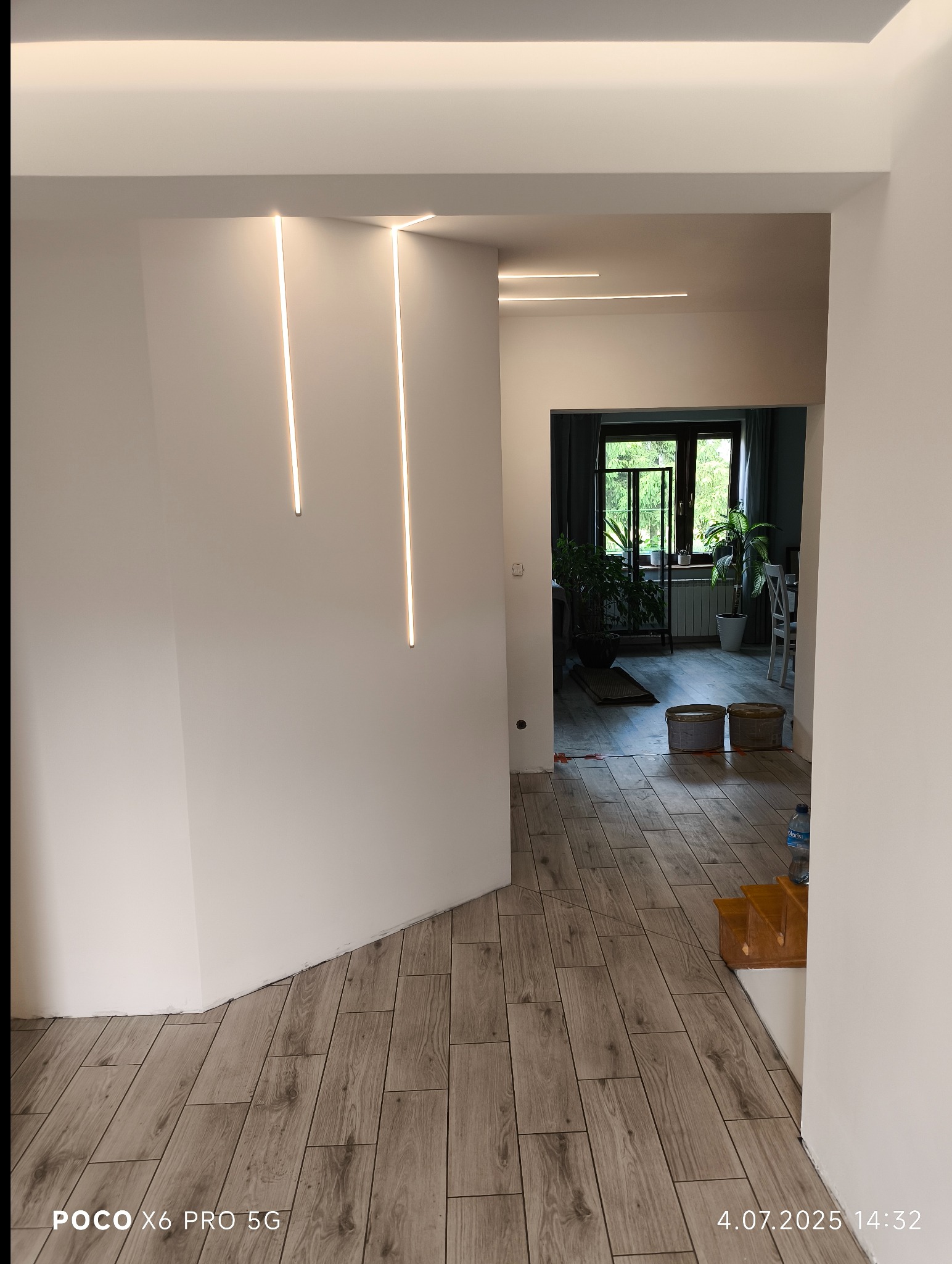 Nowoczesne wykończenie wnętrza: drewnopodobna podłoga, minimalistyczne oświetlenie LED w ścianach, widok na salon z roślinami. Detal: narożnik ściany.