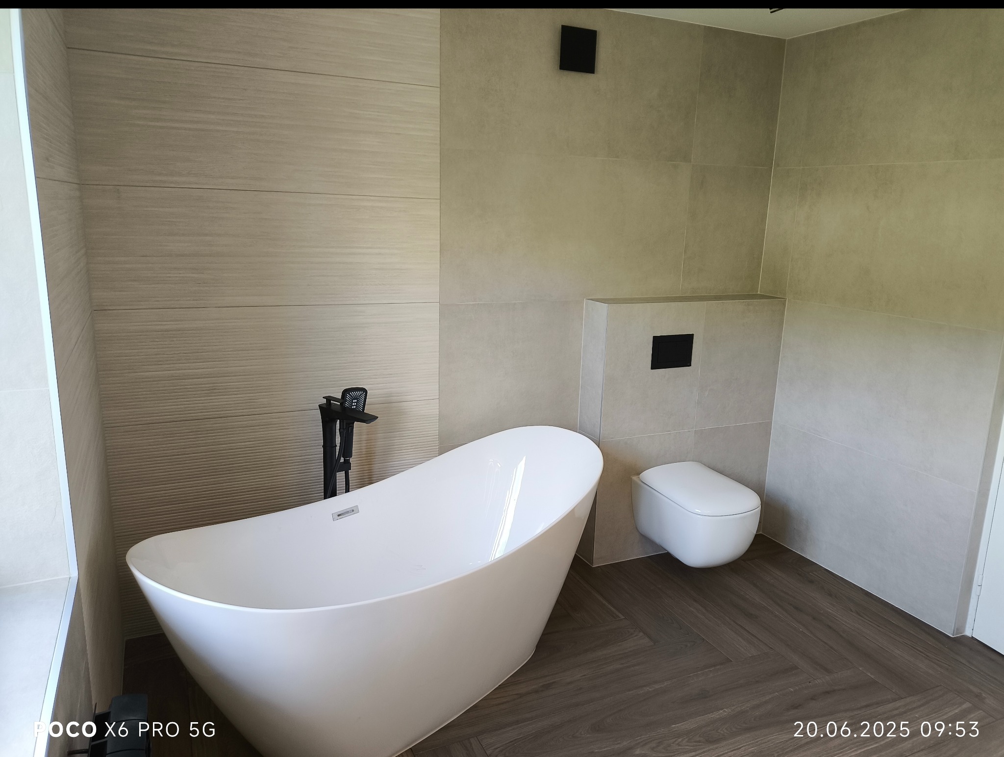 Nowoczesna łazienka z wolnostojącą wanną, podwieszaną toaletą i płytkami imitującymi drewno na podłodze. Minimalistyczny design, stonowana kolorystyka, czarna armatura.