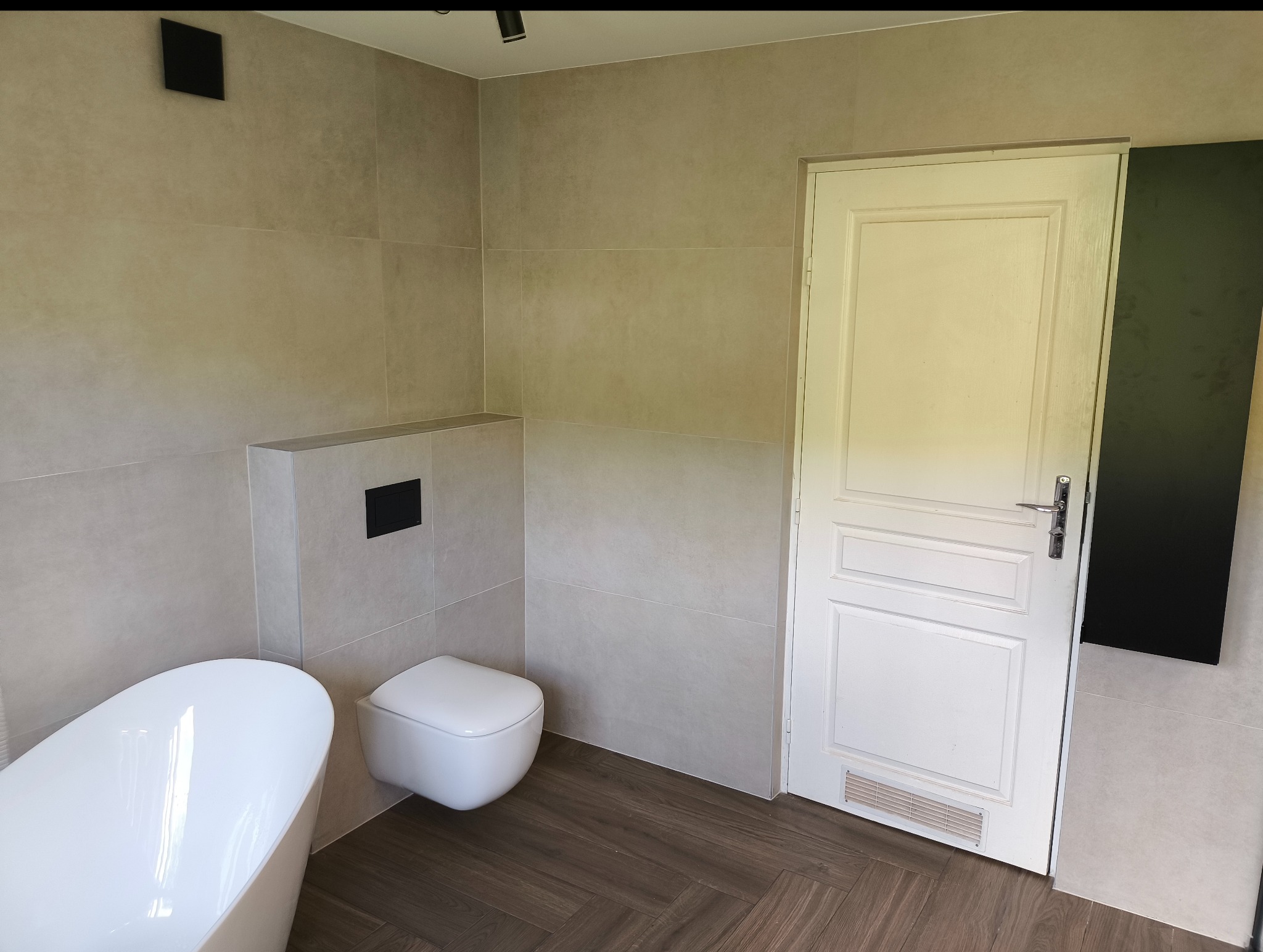 Jasna łazienka po remoncie: beżowe płytki, drewnopodobna podłoga w jodełkę, biała wanna wolnostojąca i podwieszana toaleta. Minimalistyczny design, nowoczesne oświetlenie.