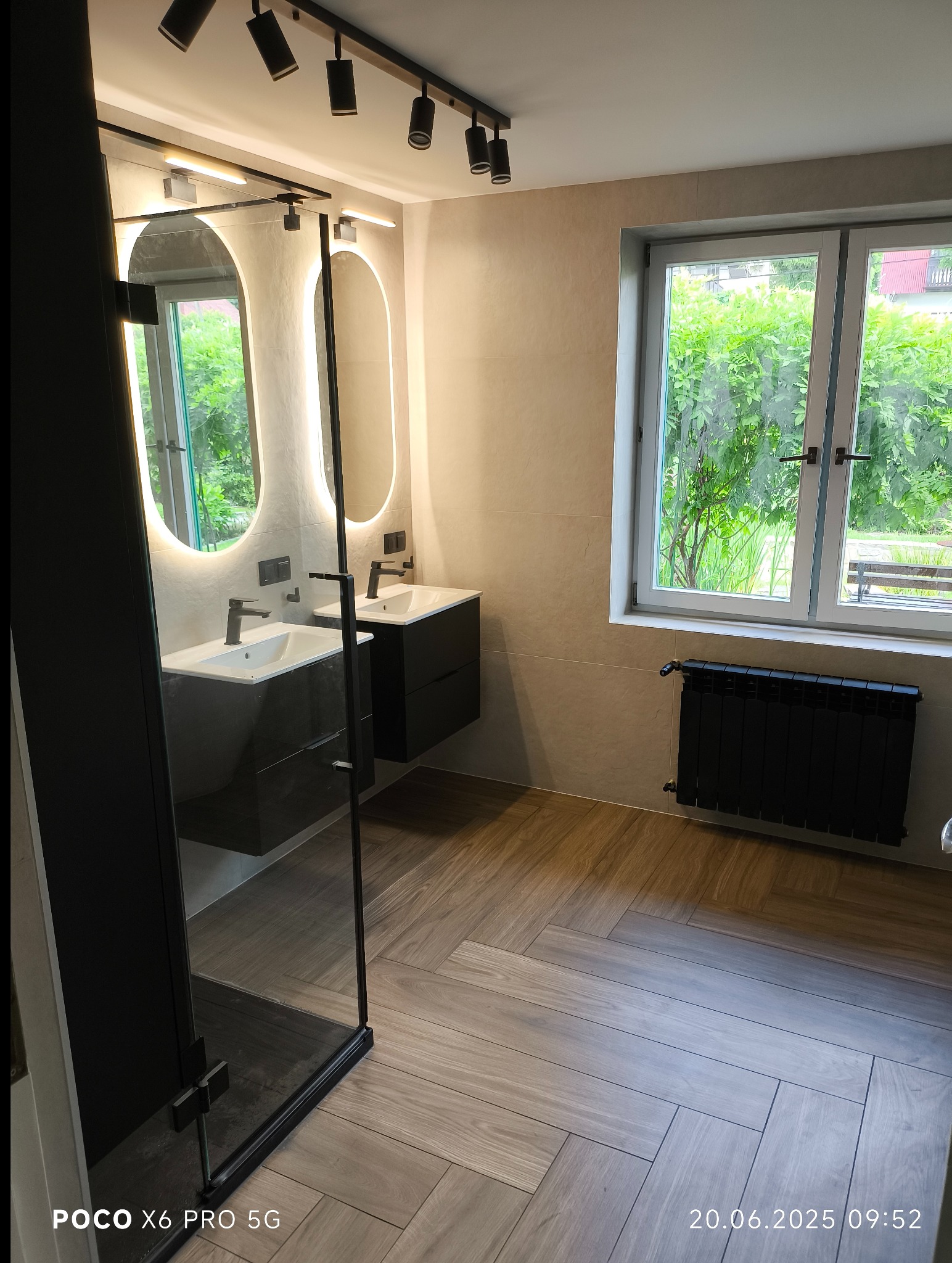 Nowoczesna łazienka z dwoma umywalkami, podświetlanymi lustrami i drewnopodobną podłogą ułożoną w jodełkę. Widoczne okno z widokiem na zieleń i czarny grzejnik.