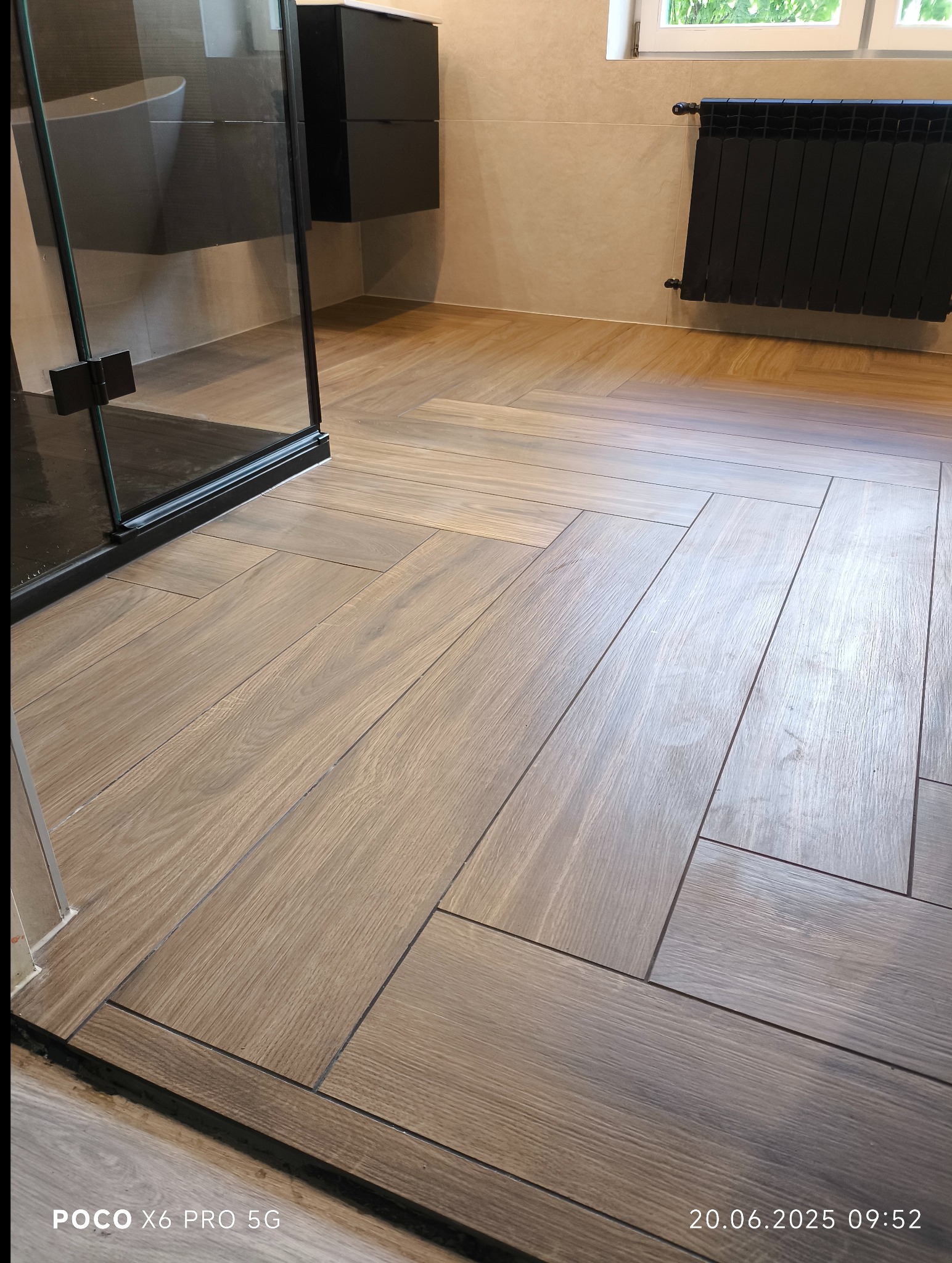 Ułożona w jodełkę podłoga z płytek imitujących drewno w łazience z kabiną prysznicową i grzejnikiem. Jasne ściany i okno w tle.