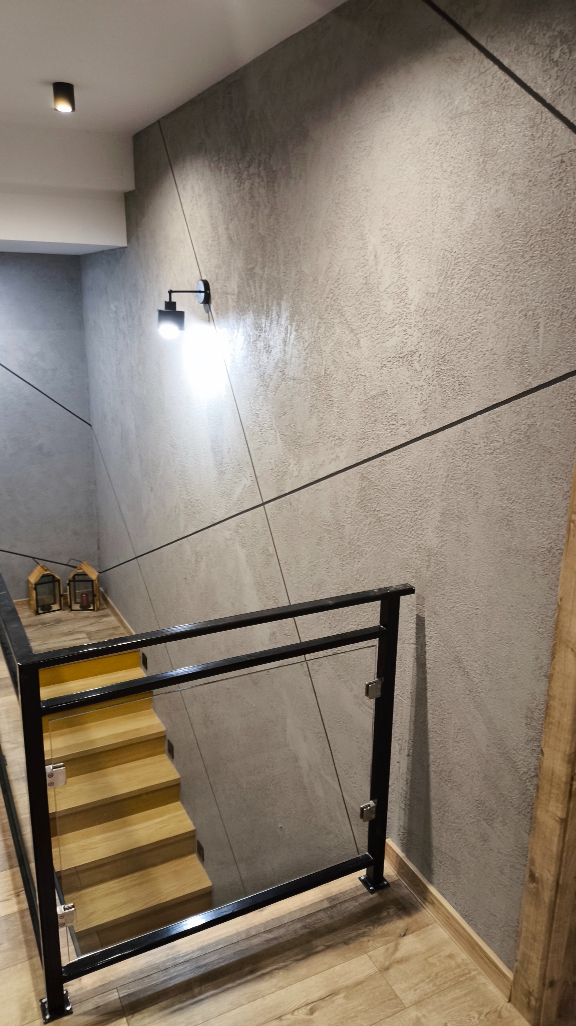 Nowoczesne wykończenie klatki schodowej z drewnianymi schodami, szarą ścianą z geometrycznym wzorem i szklaną balustradą z czarnymi elementami.