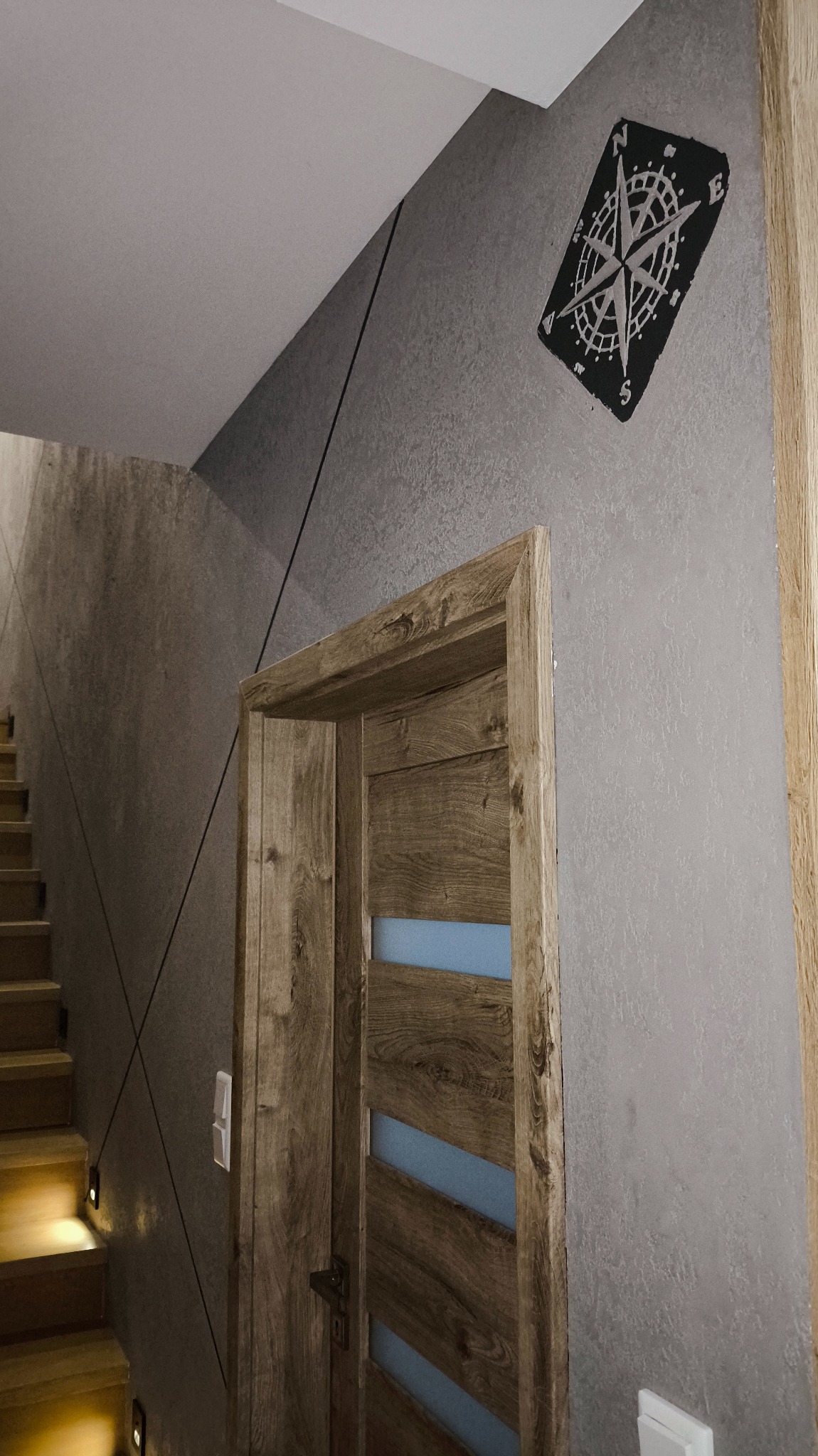 Szara ściana z dekoracyjnym kompasem i drewniane drzwi z przeszkleniami, widok na schody z oświetleniem punktowym. Nowoczesne wykończenie wnętrza.
