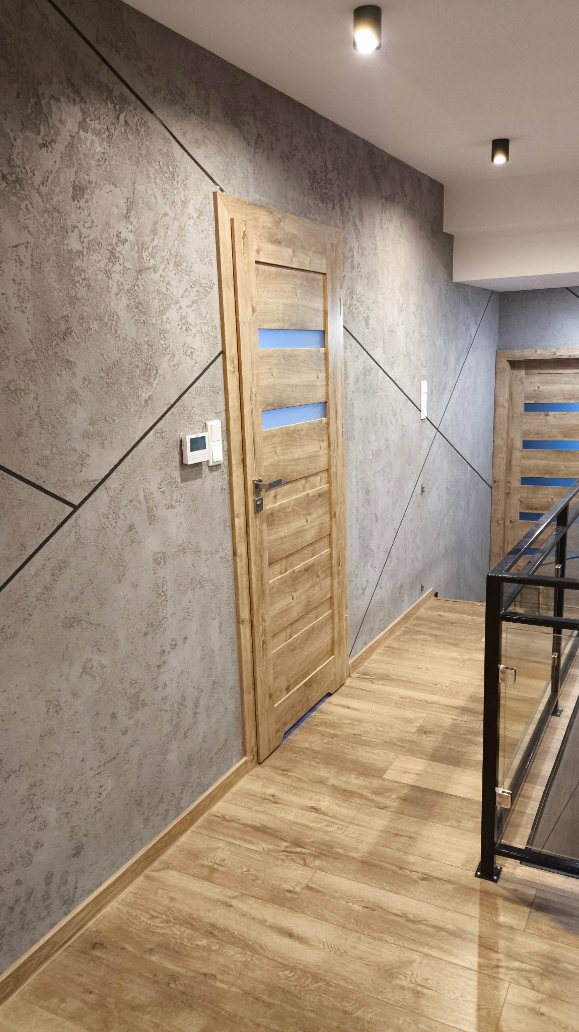 Wnętrze korytarza z dekoracyjną szarą ścianą z czarnymi liniami, drewnianą podłogą i dwiema drewnianymi drzwiami z niebieskimi akcentami. Nowoczesne oświetlenie sufitowe.